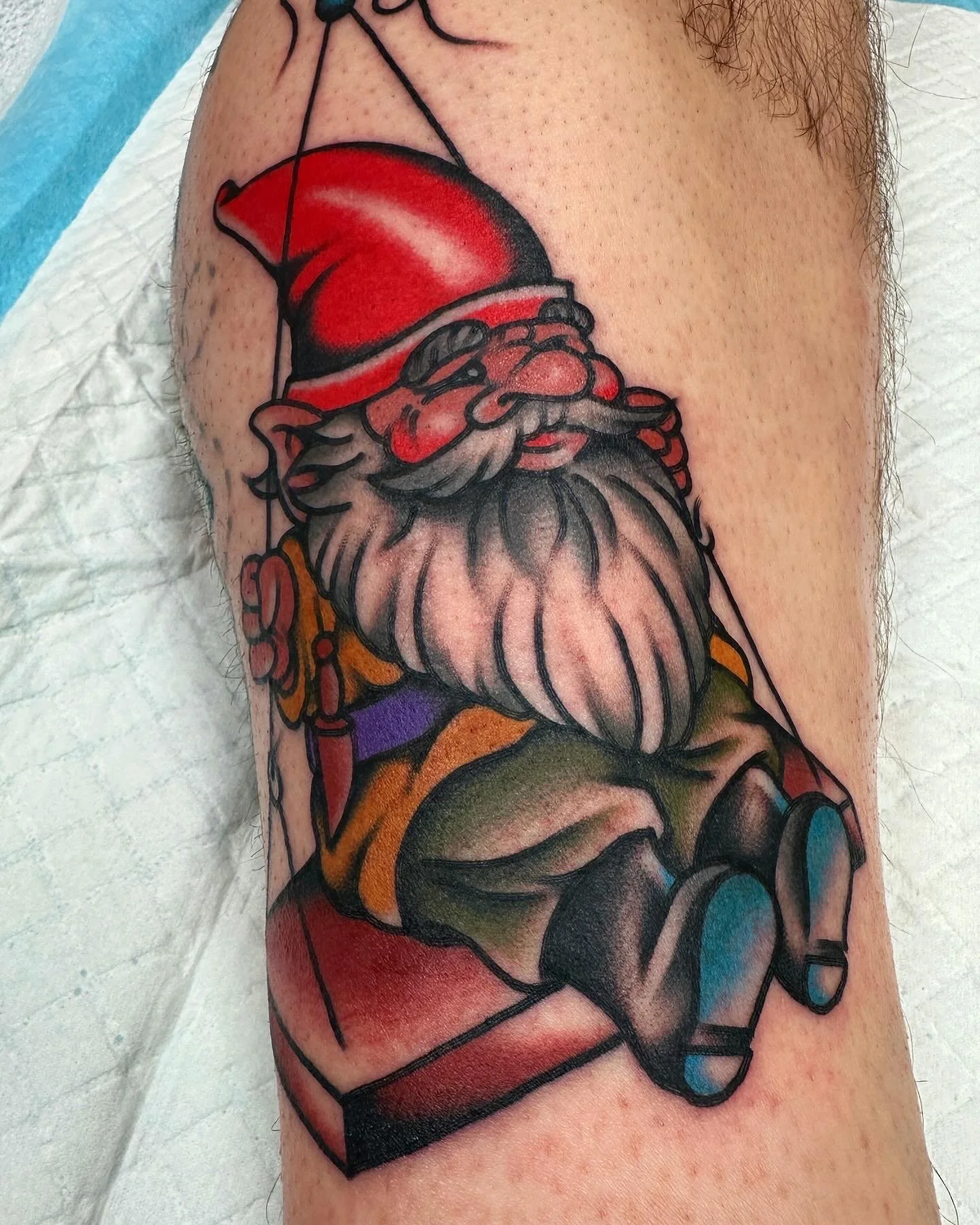 Colourful tattoo of a garden gnome with a red hat, white beard, and red cheeks on a person's upper arm.