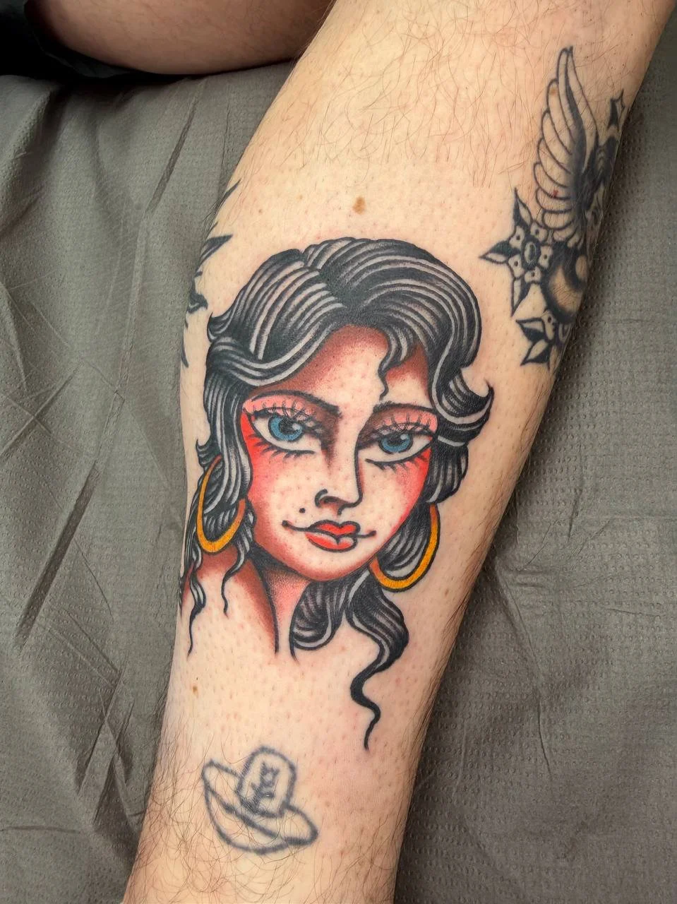 Tattoo of a woman with dark wavy hair, blue eyes, red cheeks, red lipstick, gold hoop earrings, and a beauty mark, on a person's arm. 