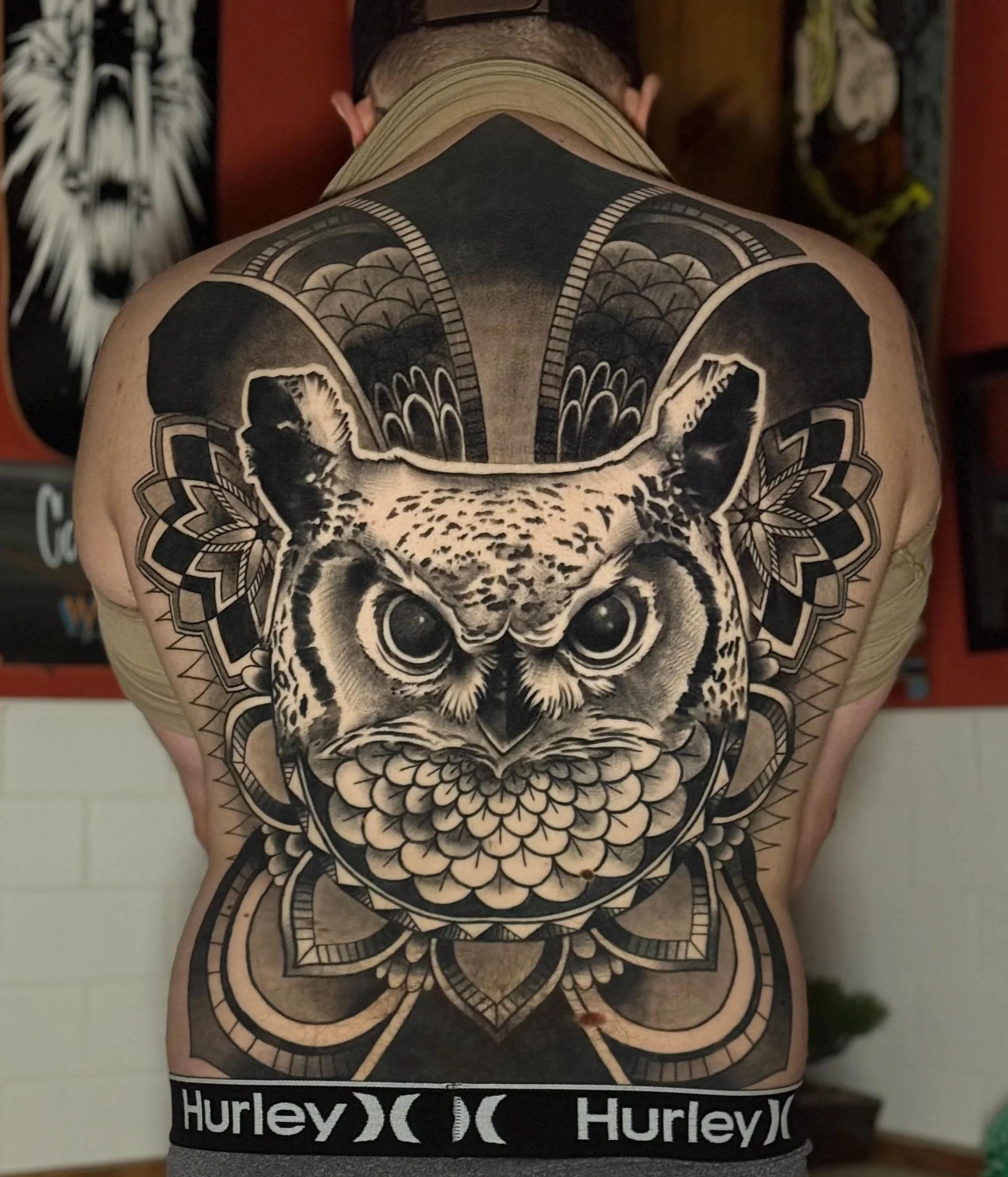 Person with a large, detailed black and grey Owl tattoo on their back. The tattoo features intricate floral and geometric patterns. Black and grey realism back piece tattoo.