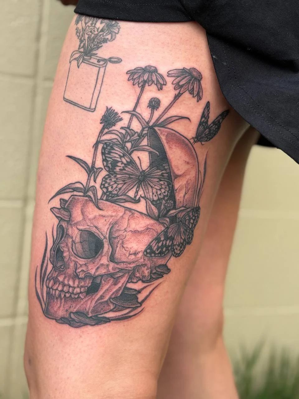 Tattoo of a skull, butterflies, flowers, and a jar on a person's thigh. Illustrative blackwork tattoo.