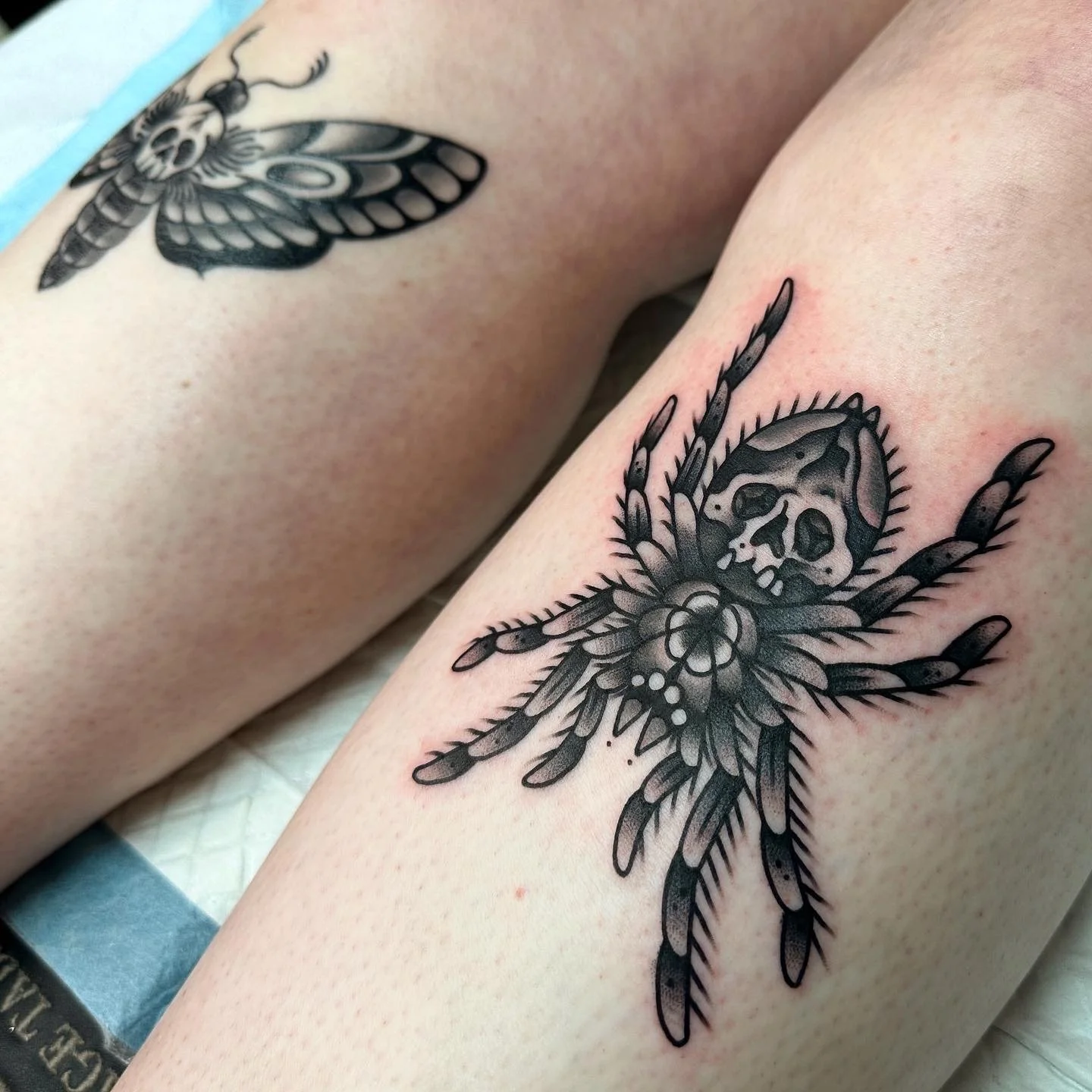 Black and gray tattoo of a spider with a skull and death moth on a person's leg.