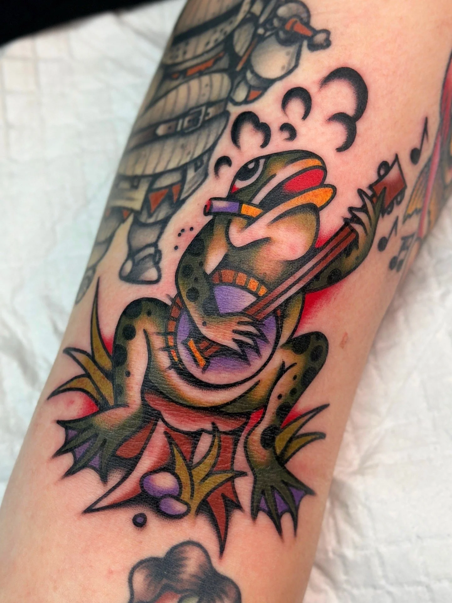 Colourful tattoo of a cartoon-style frog playing a banjo on a person's arm. Bold American traditional.