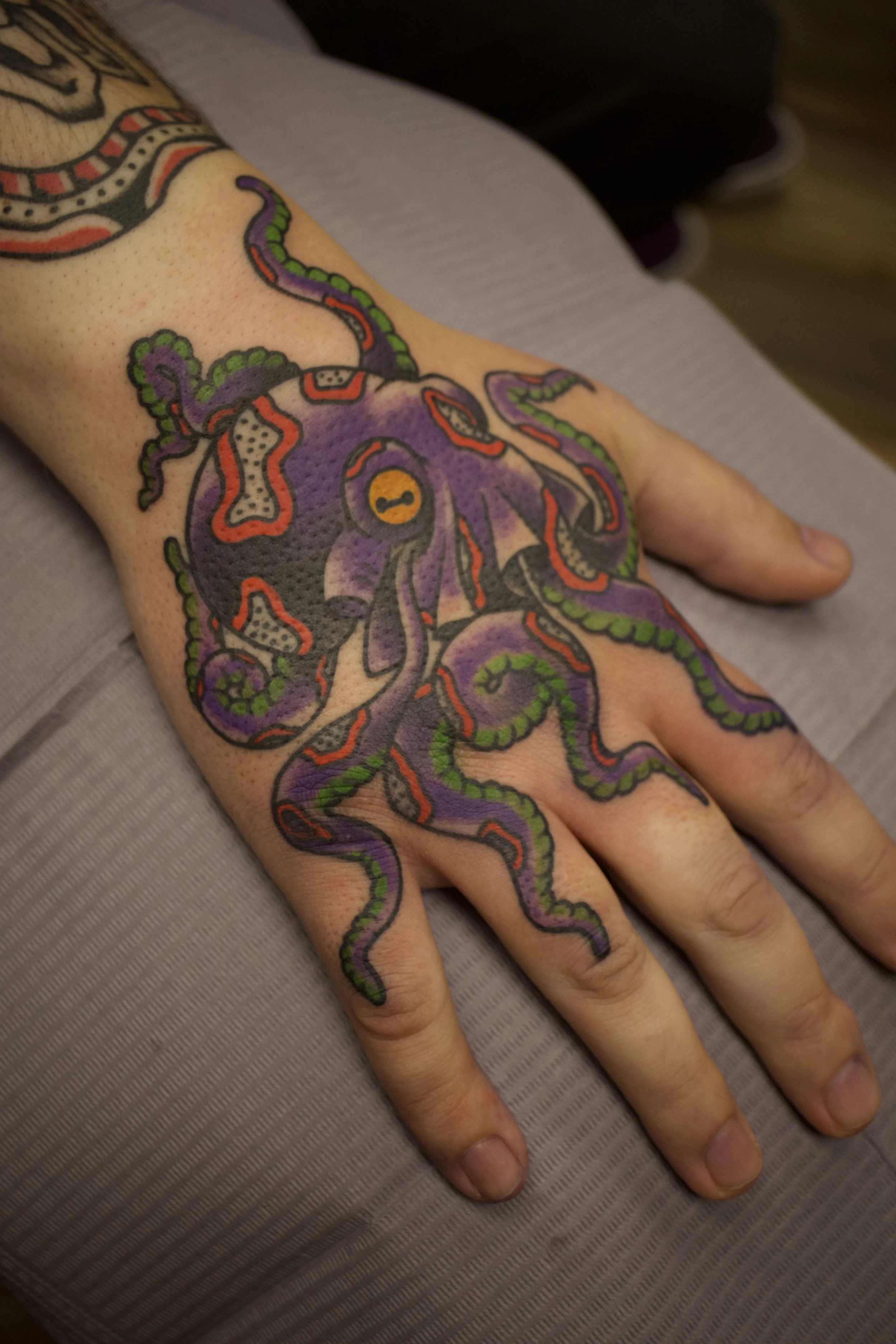 A hand with colorful tattoo art of an octopus covering the back of the hand and fingers, featuring purple, green, red, black, and yellow ink details.