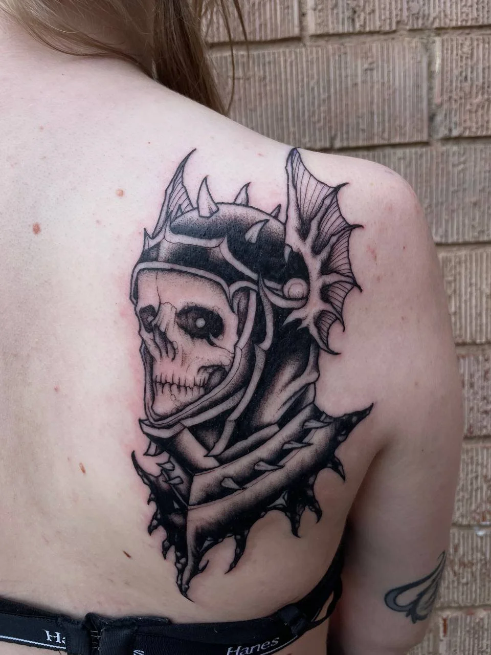 Black and gray tattoo of a skull with a knight helmet and armour, surrounded by thorny, bat-like wings, on helm.