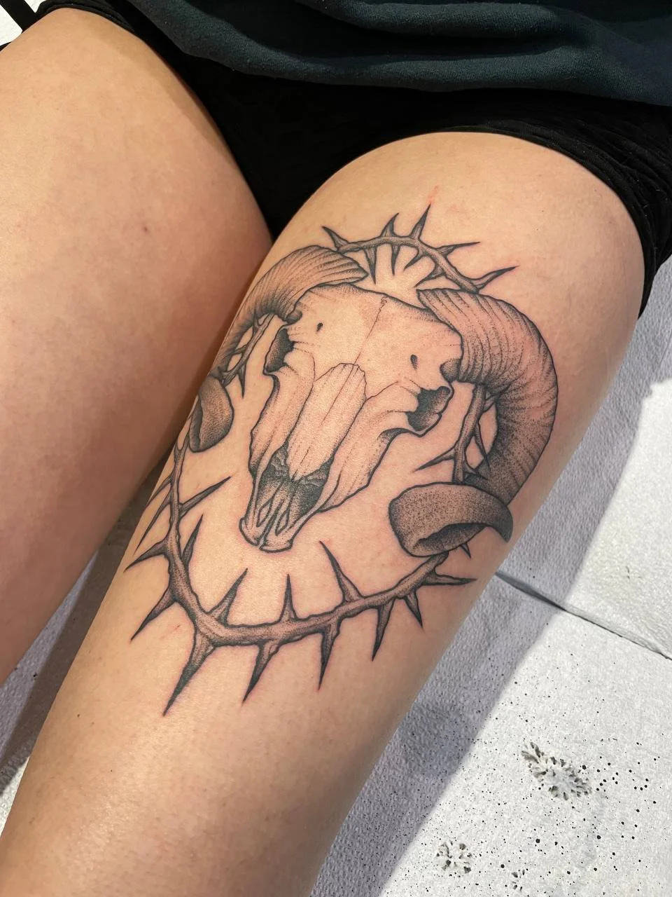 Tattoo of a ram's skull with large curved horns, surrounded by thorny vines and a halo of spikes, on a person's thigh.