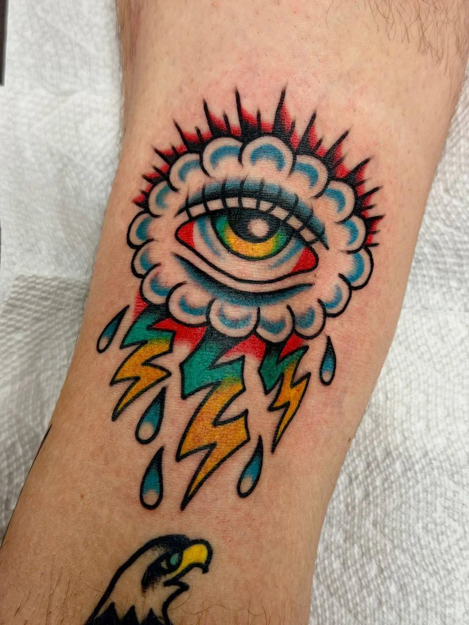 Colorful tattoo of a sinister eye inside a cloud, with rain droplets and lightning bolts underneath, on a person's arm. Eye of the storm tattoo. Bold American traditional tattoo.