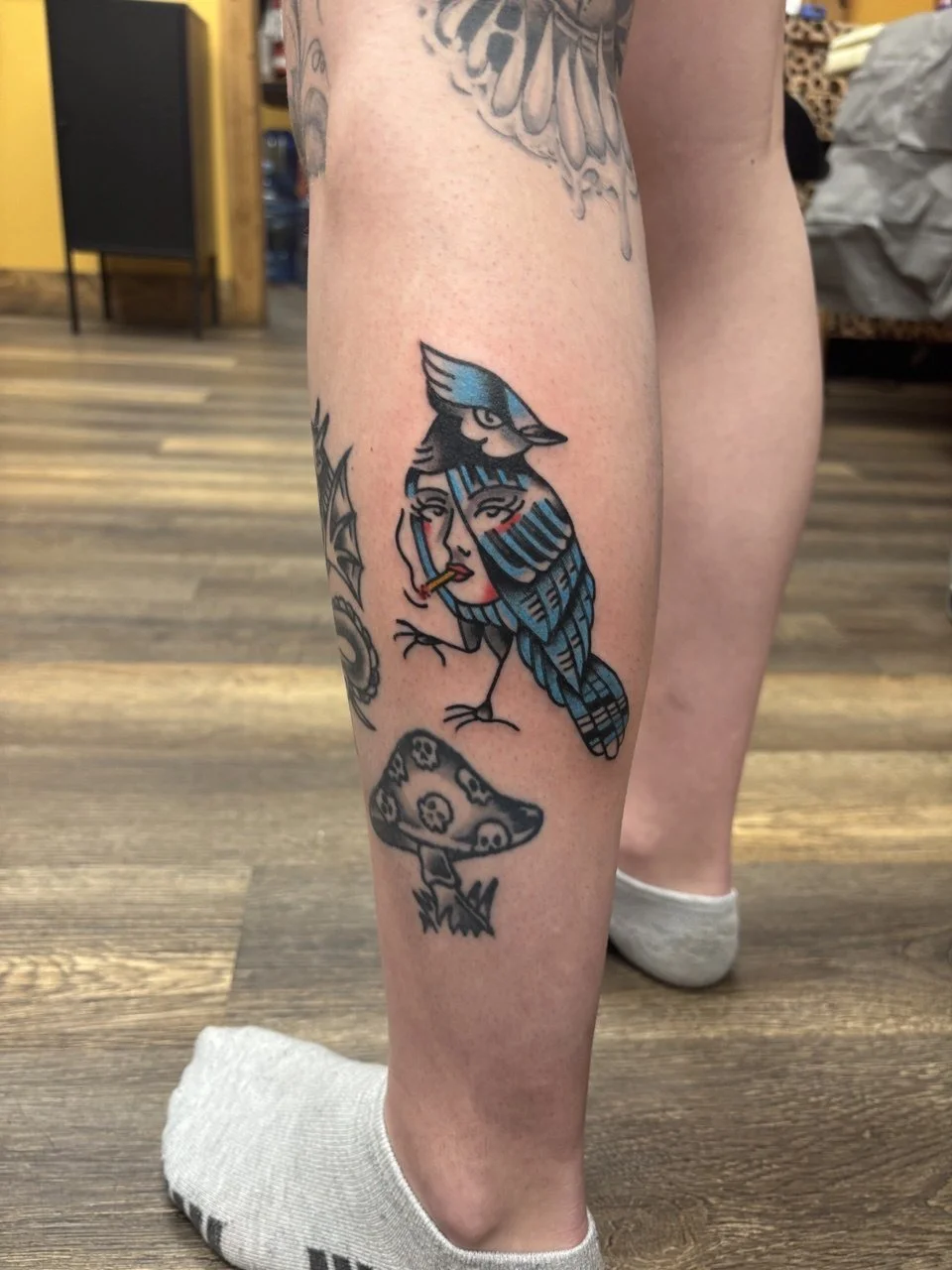 Tattoo of a bluejay with a woman smoking inside the design, on person's calf. Full colour American traditional tattoo. 