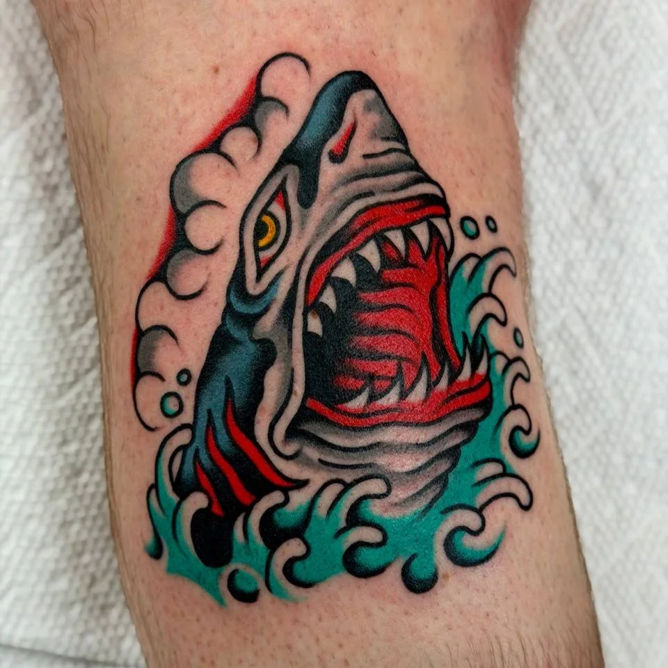A colourful tattoo of a ferocious shark with its mouth open, surrounded by water and waves. Bold American traditional tattoo.