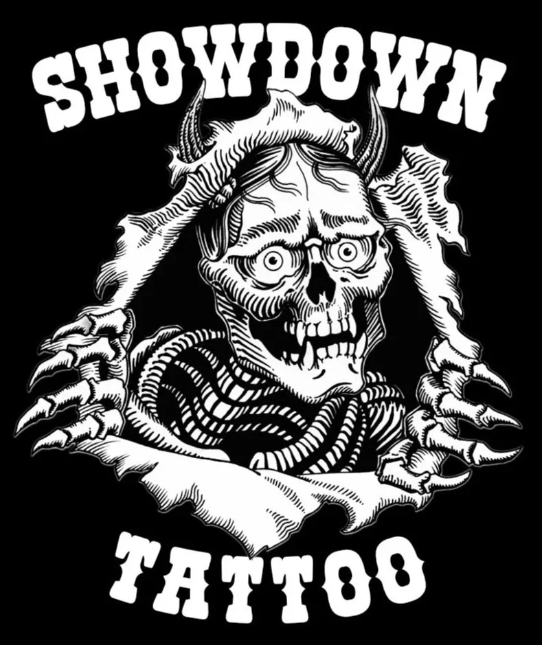 Black and white graphic of a skeleton with horns and a skull face, holding a torn banner, with the words "SHOWDOWN TATTOO" in bold text above and below. Showdown Tattoo "RIPPER" logo.