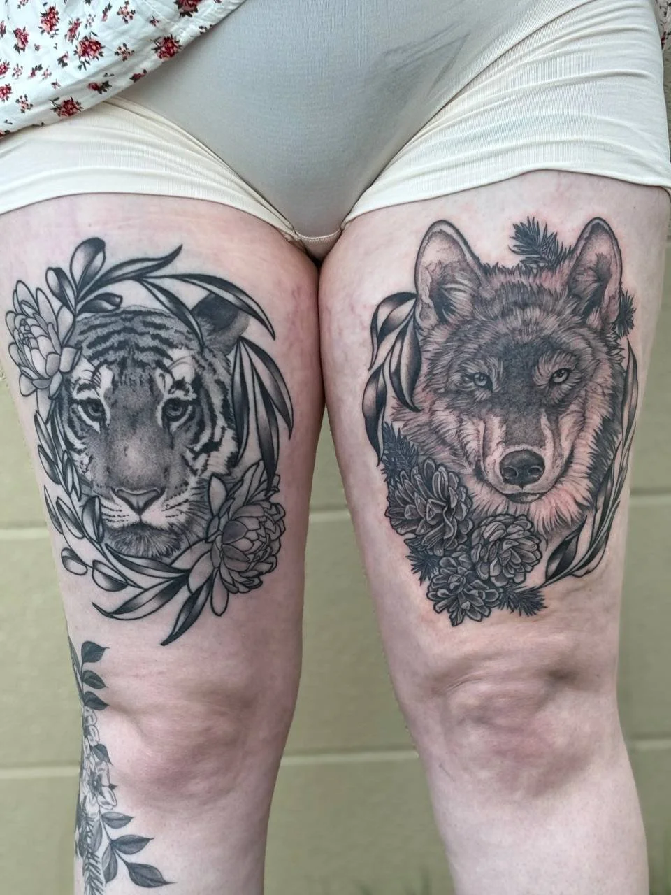 Tattoo of a tiger on the left thigh and a wolf on the right thigh surrounded by floral designs. Illustrative blackwork designed tattoo.