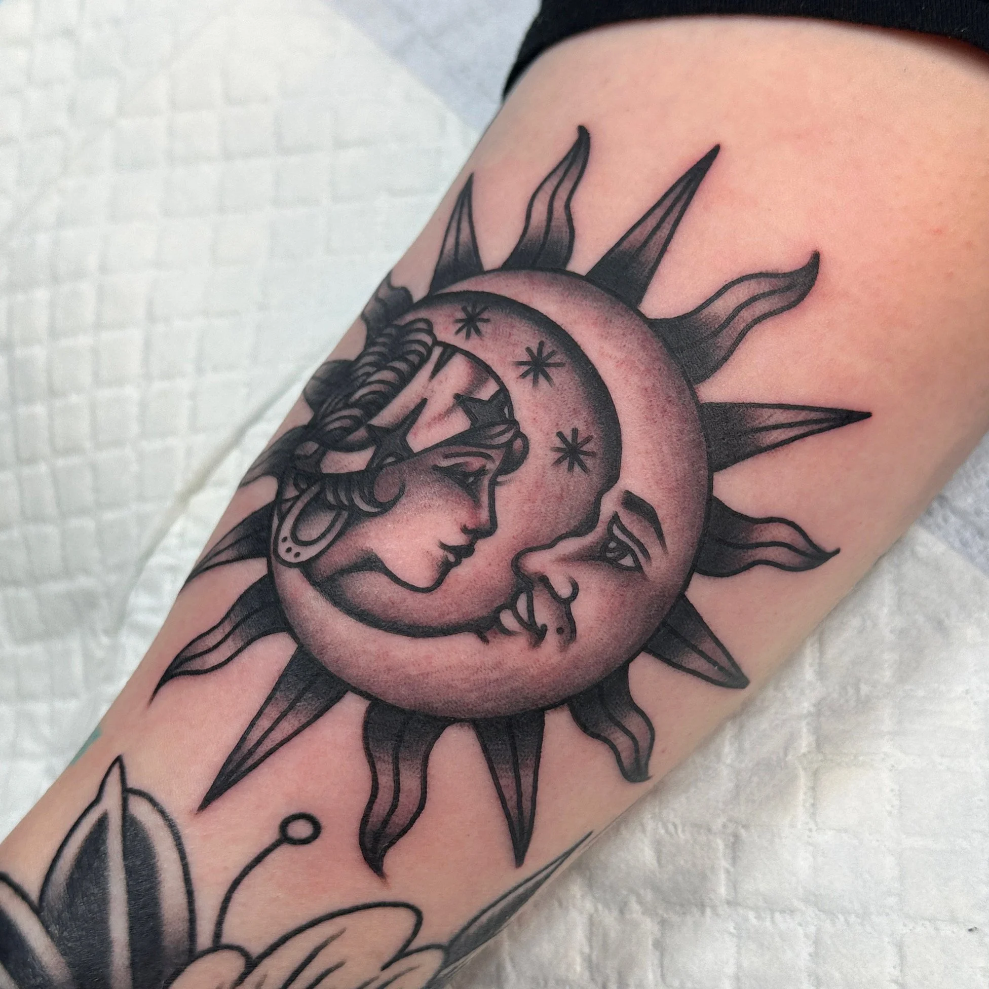 Close-up of a person's arm with a tattoo of a moon and woman facing eachother, surrounded by lines and small stars.