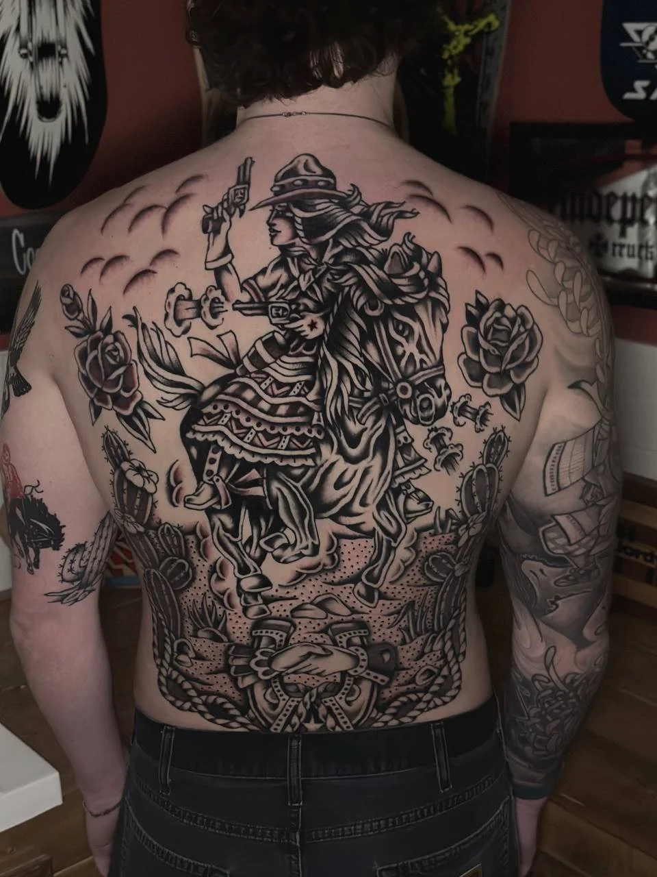 Tattoo of a mounted cowgirl with a gun, surrounded by roses and decorative elements covering the back and arms. Black and grey American traditional back piece tattoo.