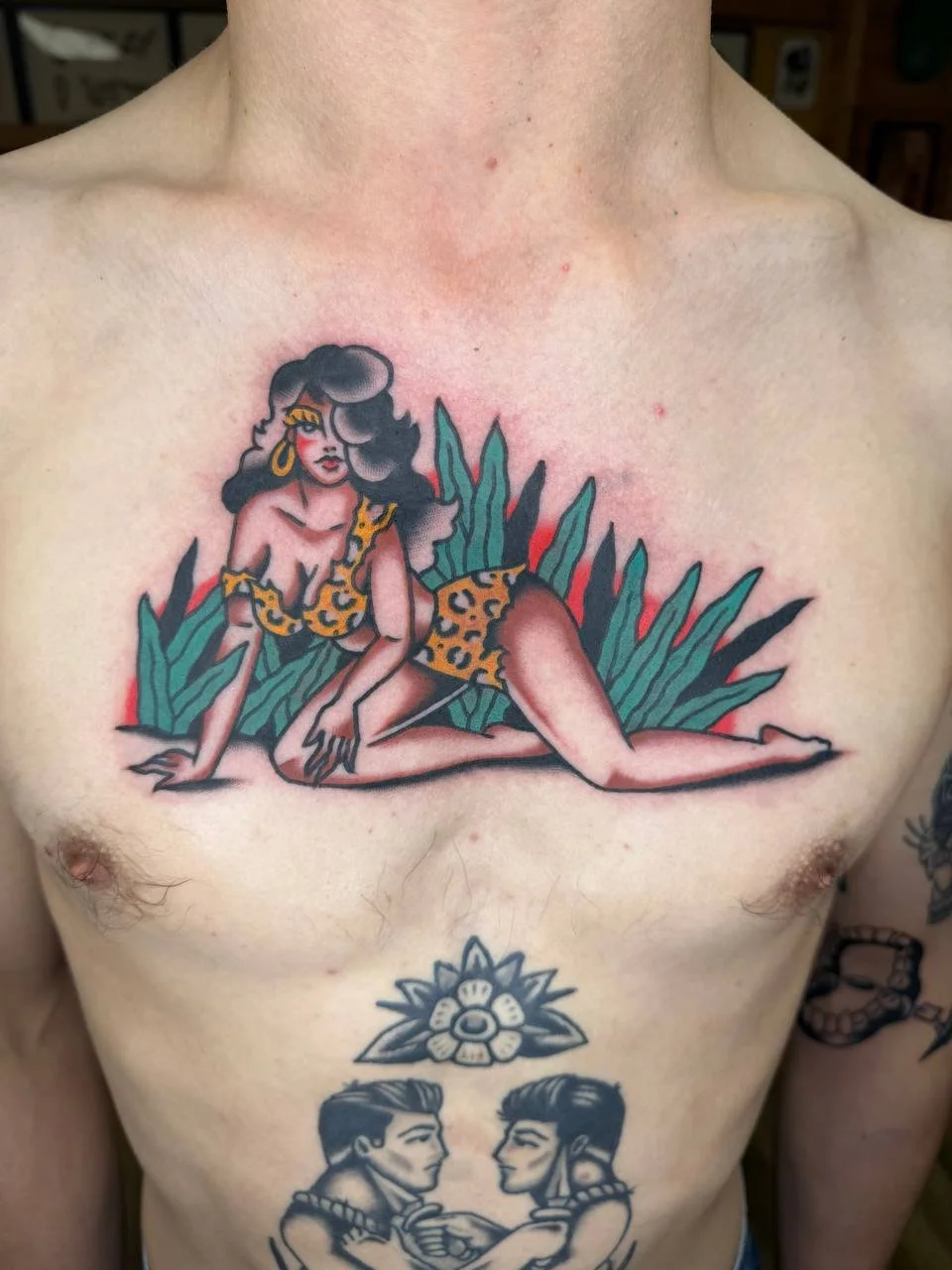Tattoo of a woman in a leopard print outfit laying on green leaves on the chest of a person. Bold American traditional chest tattoo.