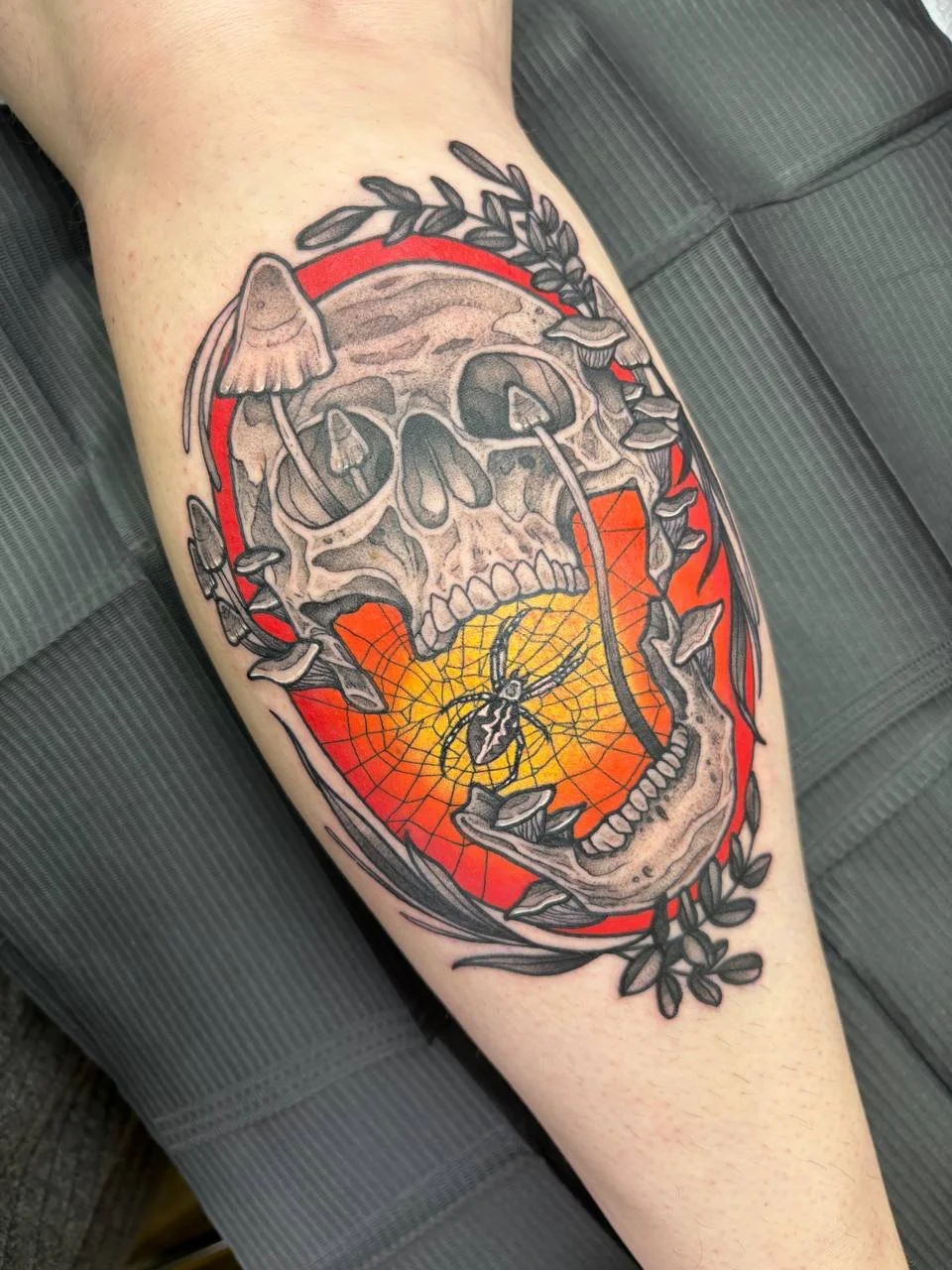 Tattoo of a human skull with an open mouth, surrounded by a wreath of leaves, with honeycomb and a spider inside the mouth, and a spider on a web in the lower part of the open mouth, with red and orange background colours.