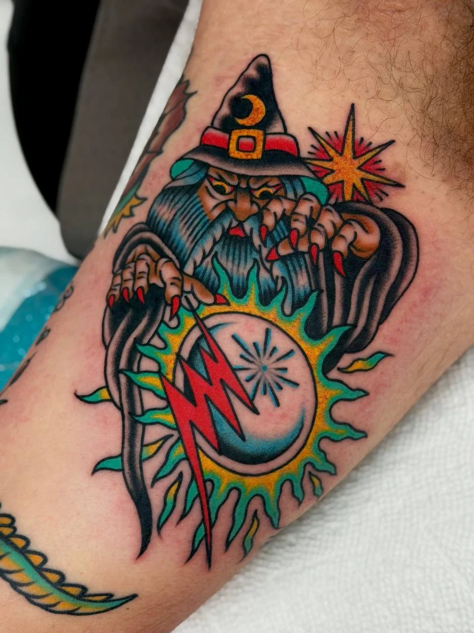 Tattoo of a wizard with a hat, long beard, and clawed hands casting a spell with a glowing orb and lightning bolts. Bold American traditional tattoo.