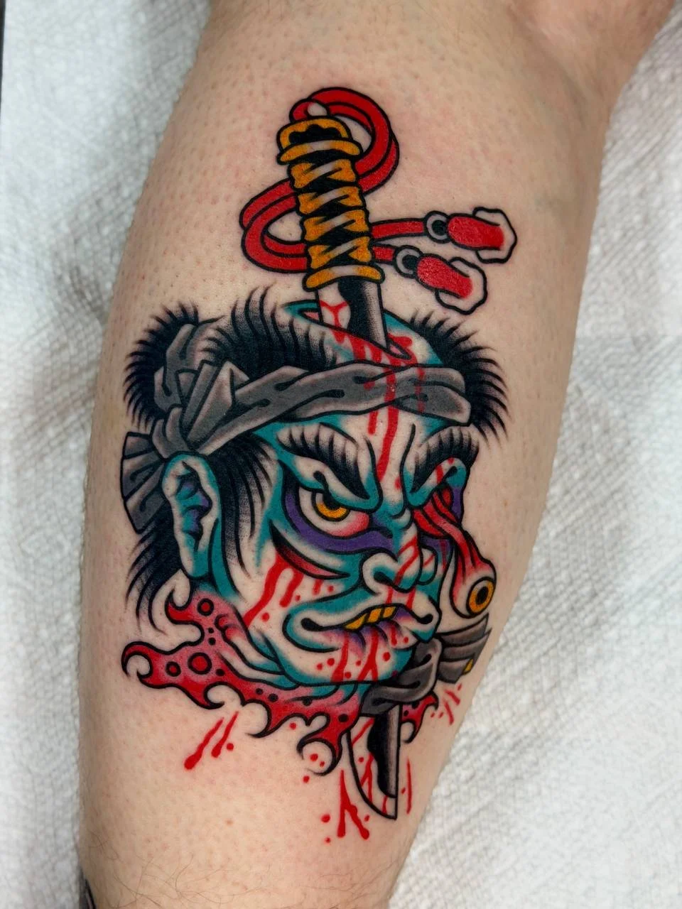 Tattoo of a traditional Japanese-style nabakubi with blood, stabbed by a sword, with a sword going through the top of the head, all in bright colours. Bold Japanese traditional tattoo.