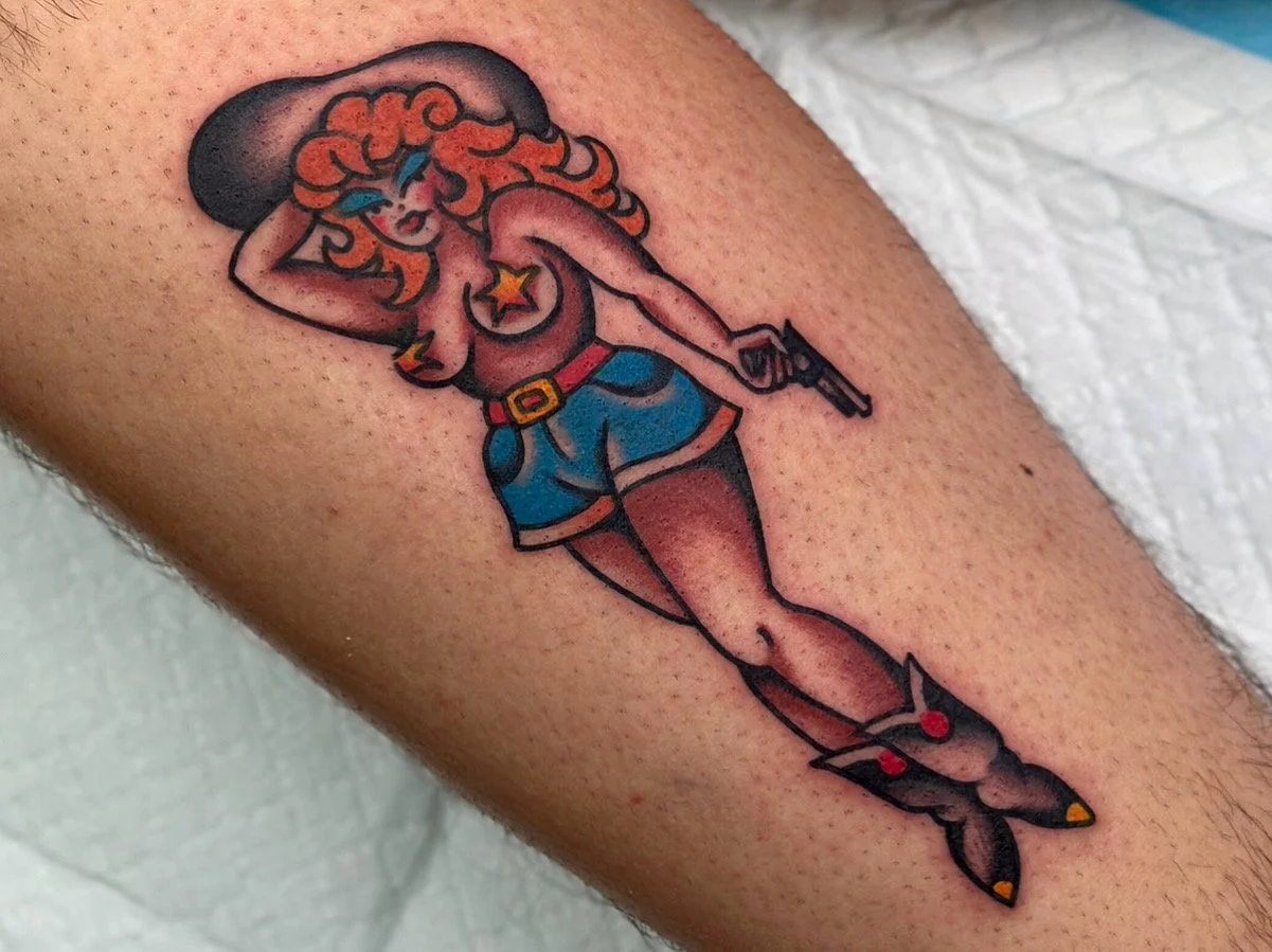 Tattoo of a american traditional pinup cowgirl with red hair, wearing a black hat, blue shorts, and a star-shaped jewelry, holding a gun.