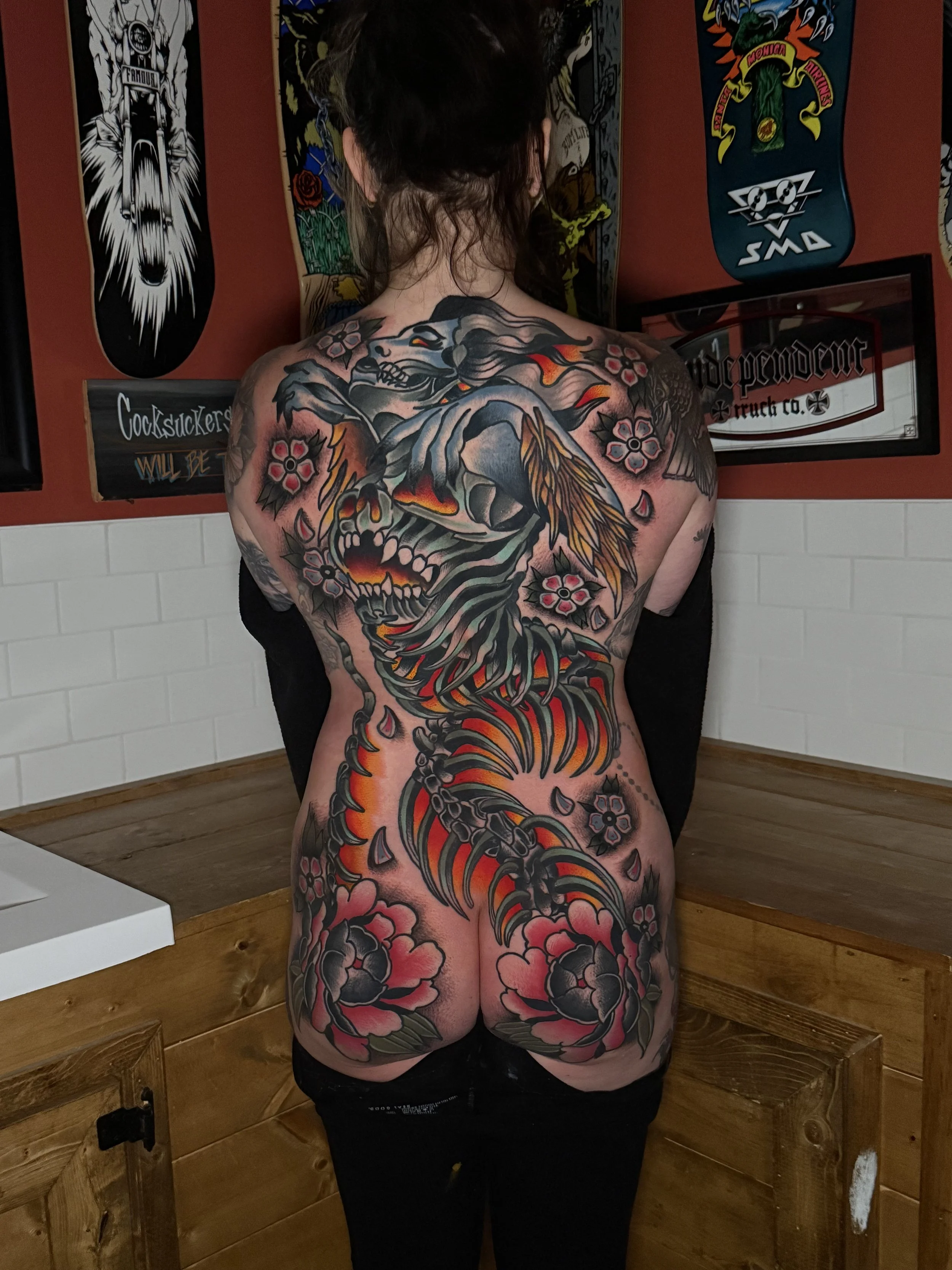 A woman with a large tattoo covering her back and extending down her arms and legs, featuring a stylized mermaid and floral patterns in black, red, yellow, and pink. Full colour back piece.
