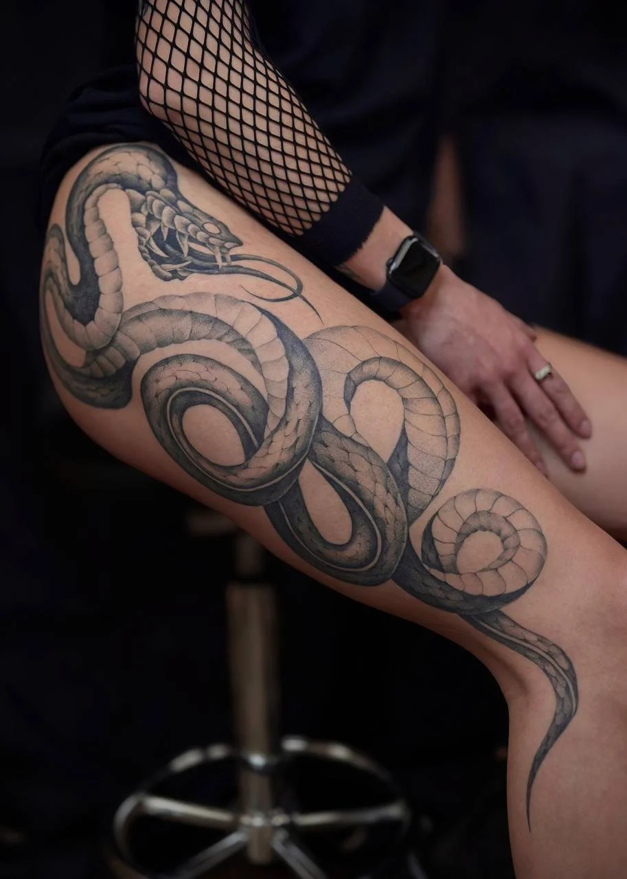 Close-up of a person's leg with a detailed black and gray tattoo of a coiled snake. Blackwork tattoo.