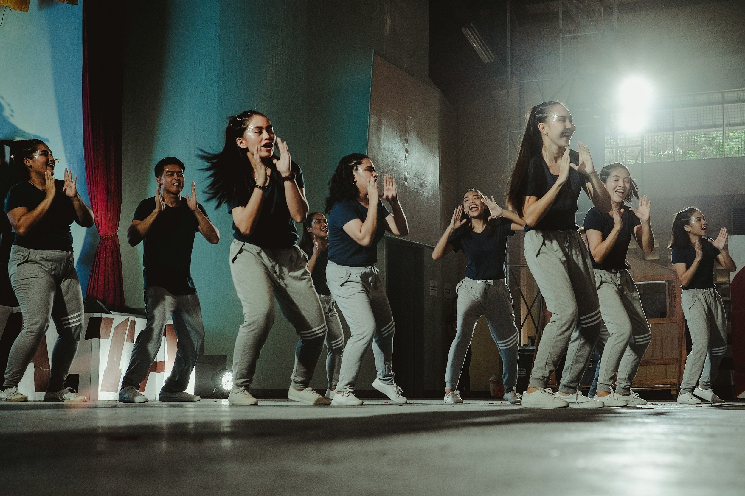 Group of young women performing a dance on stage, dressed in matching black shirts and light-colored pants, with sunlight shining through the windows in the background.