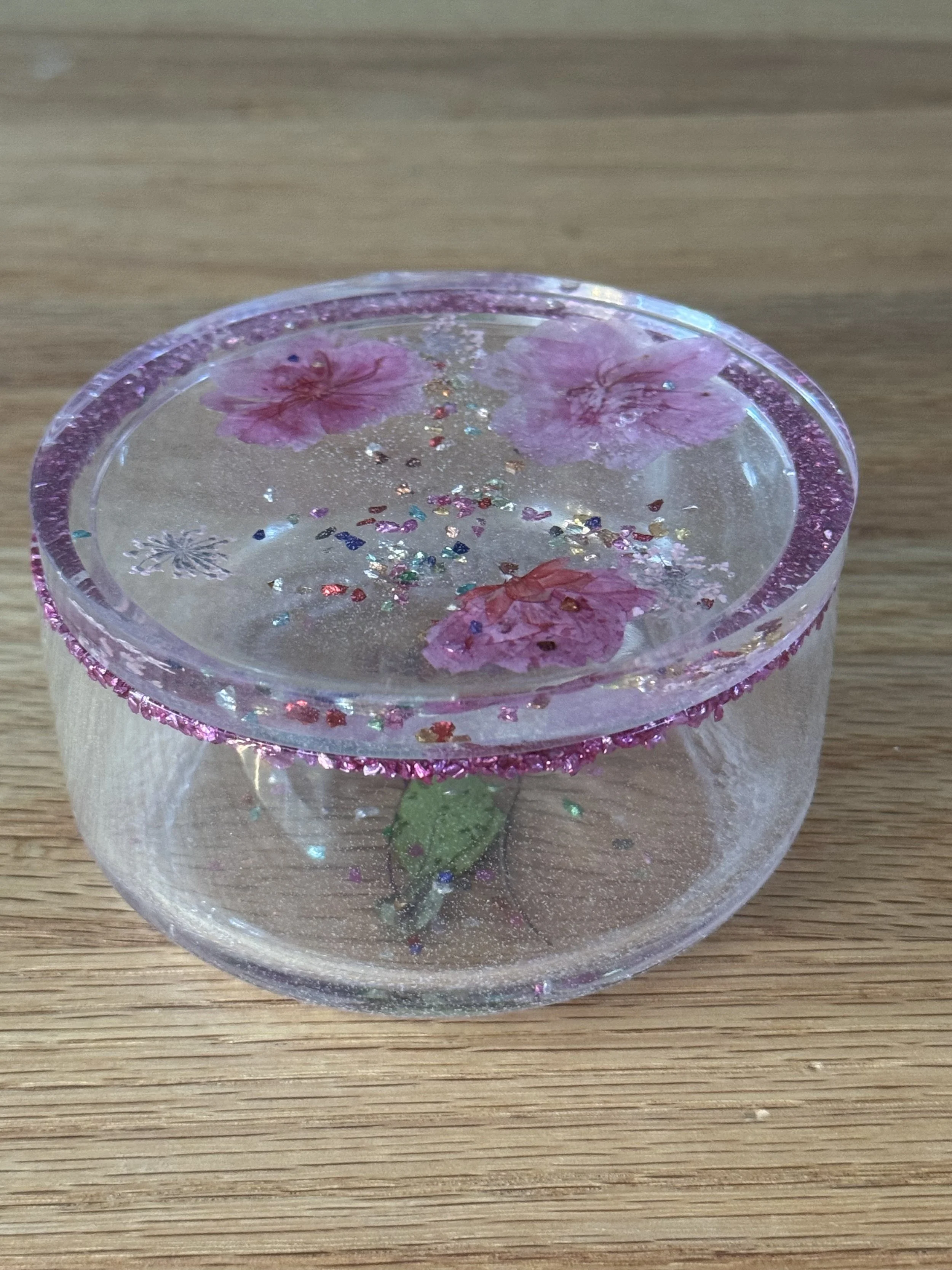 A clear glass container with pink glitter and dried pink flowers submerged inside, resting on a wooden surface.