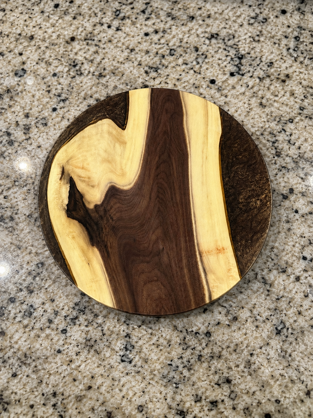 A round wooden decorative plate with a natural wood grain pattern, placed on a speckled granite countertop.