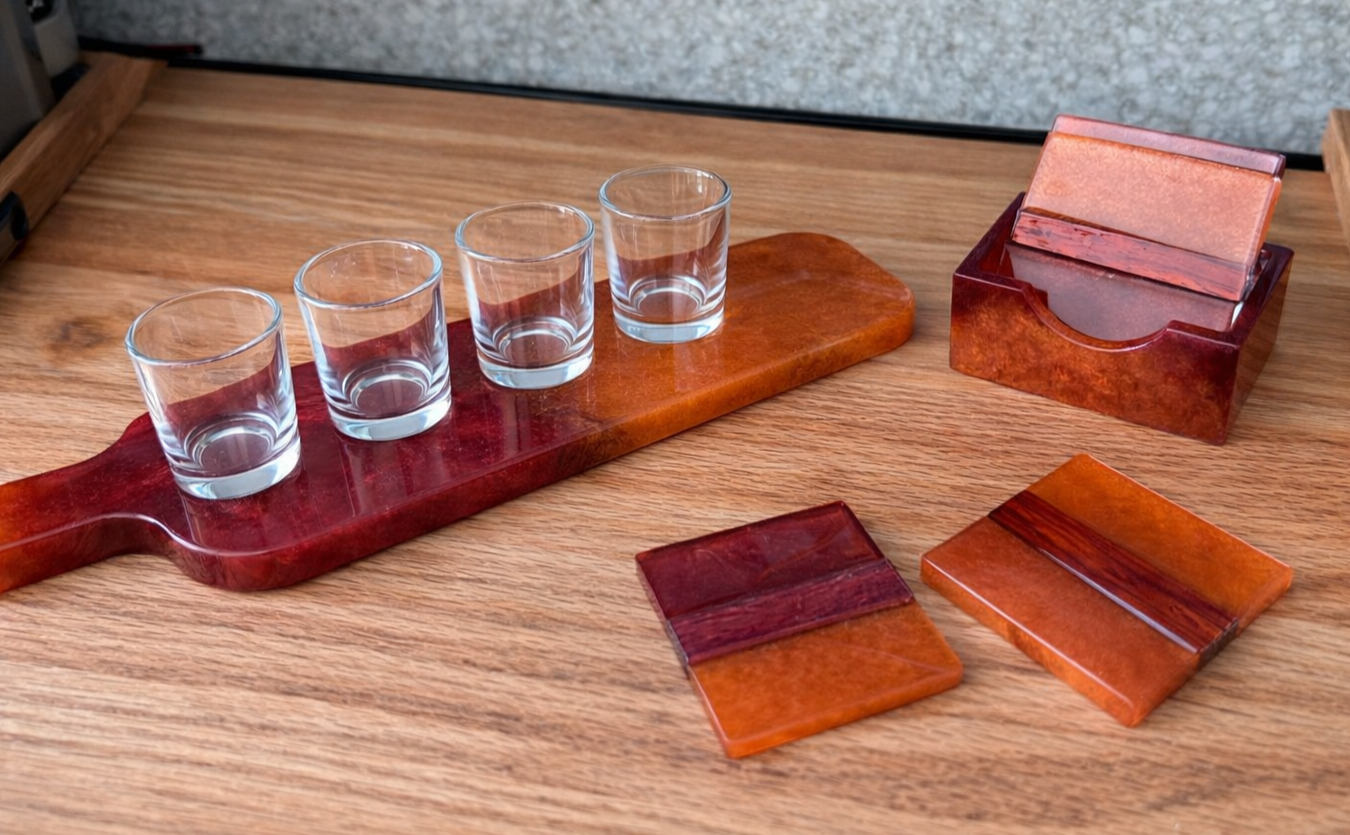 Set of four small glasses on a wooden tray, a clear glass holder with a bar of mica or similar stone, and two decorative square coasters on a wooden table.