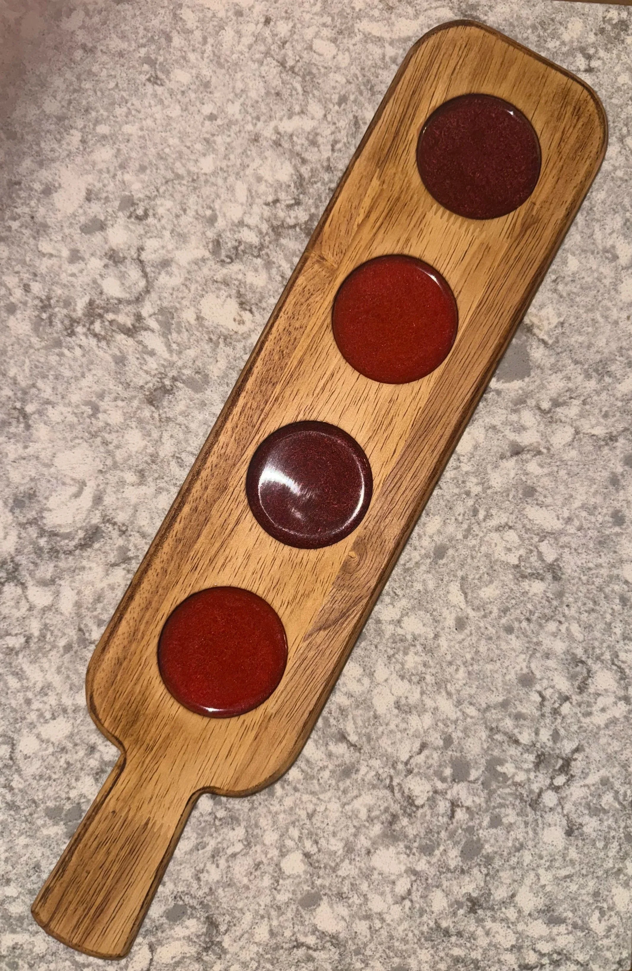 A wooden paddle with four circular depressions containing red sauces, placed on a speckled gray countertop.