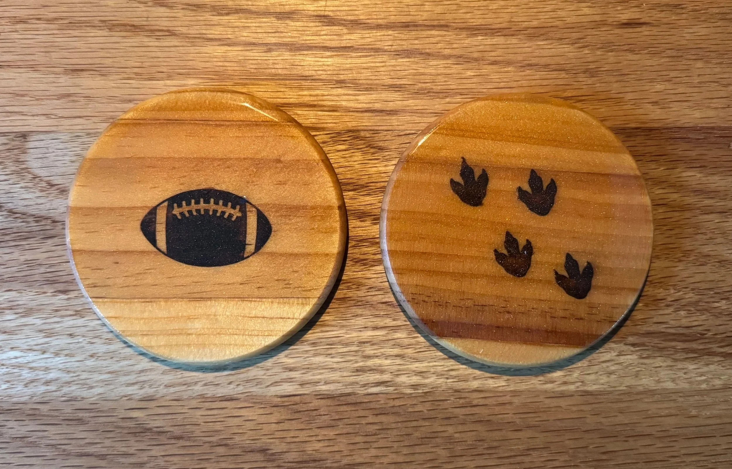 Two round wooden coasters on a wooden surface. The left coaster has a black football design, and the right coaster has black paw prints.