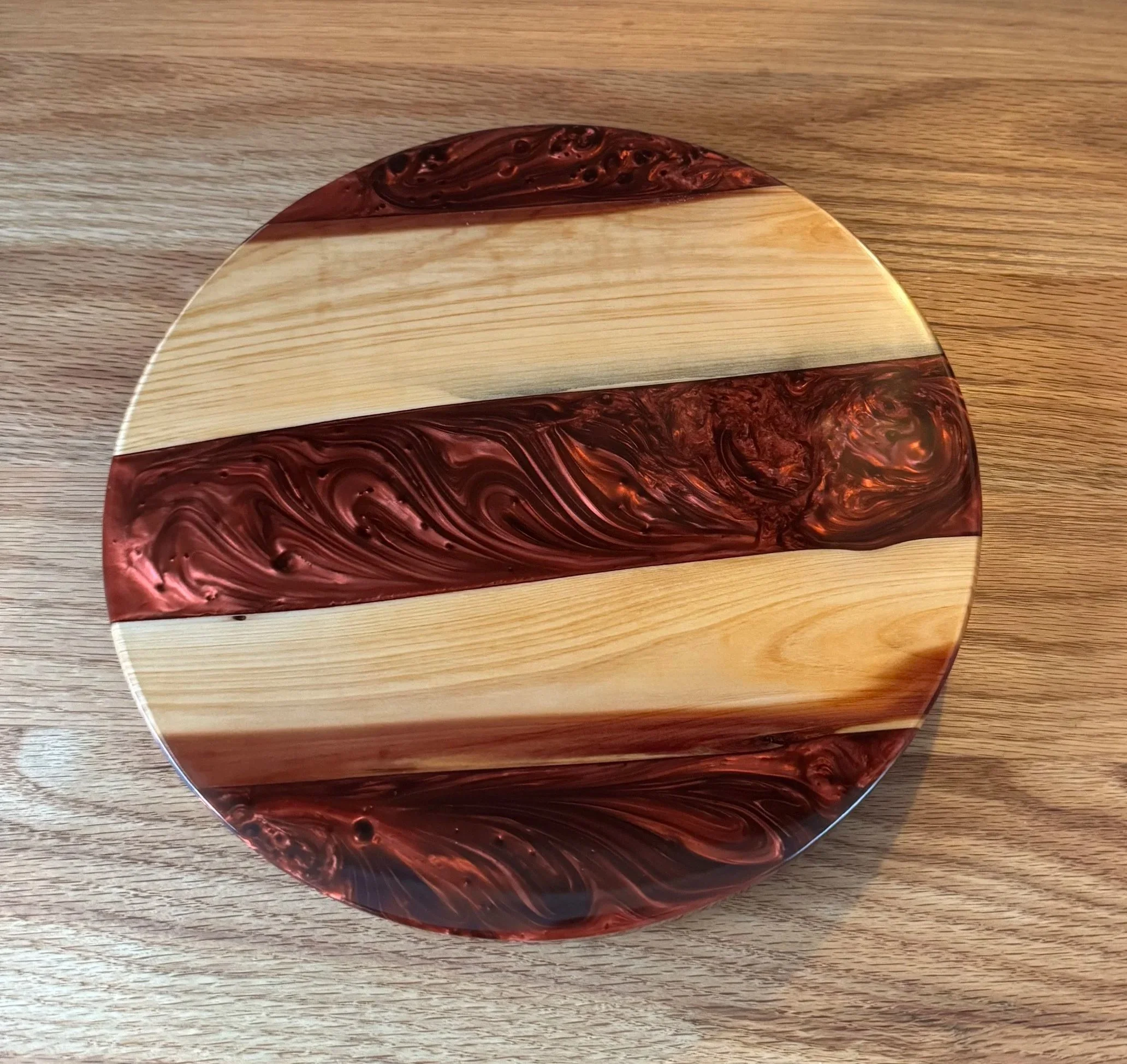 A round wooden object, possibly a decorative piece, with a mix of light natural wood and darker, swirling red and black patterns, resting on a wooden surface.
