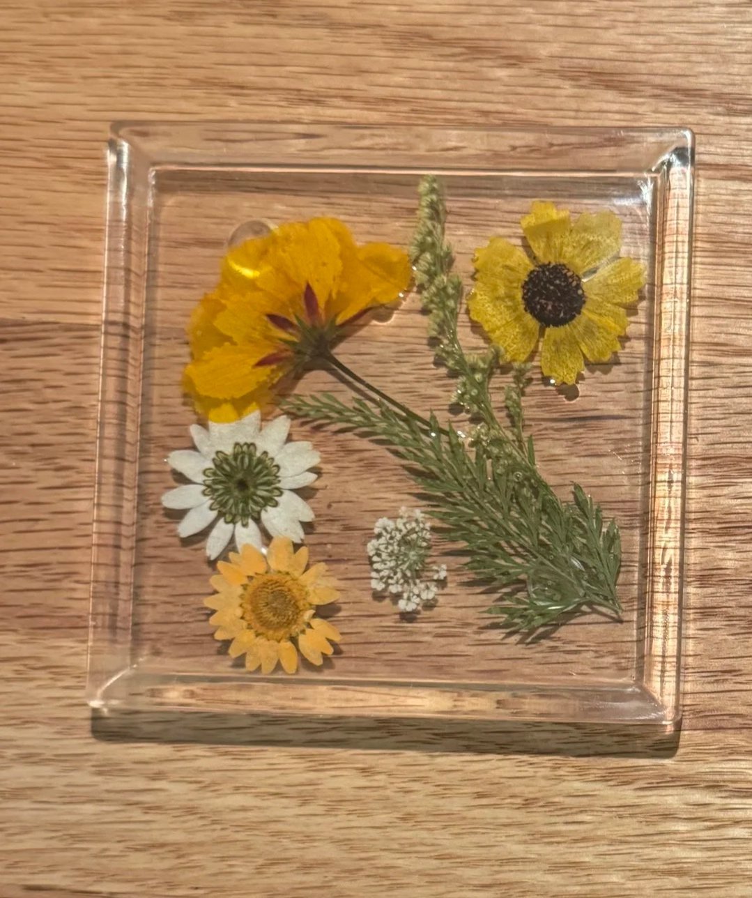Pressed yellow, white, and small white flowers with greenery in a glass container on a wooden surface.