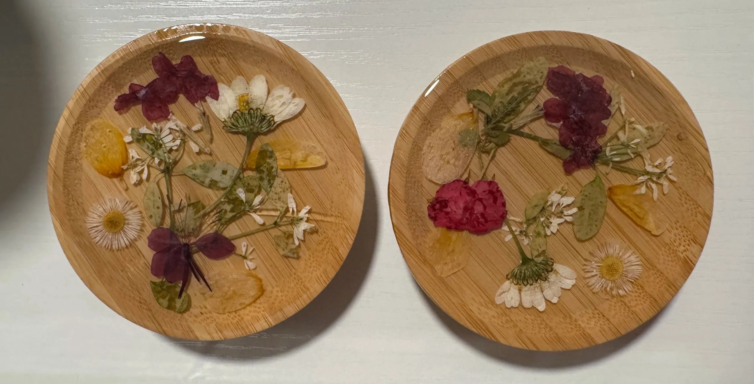 Two round wooden plates decorated with pressed flowers, leaves, and dried herbs, arranged in a floral pattern.