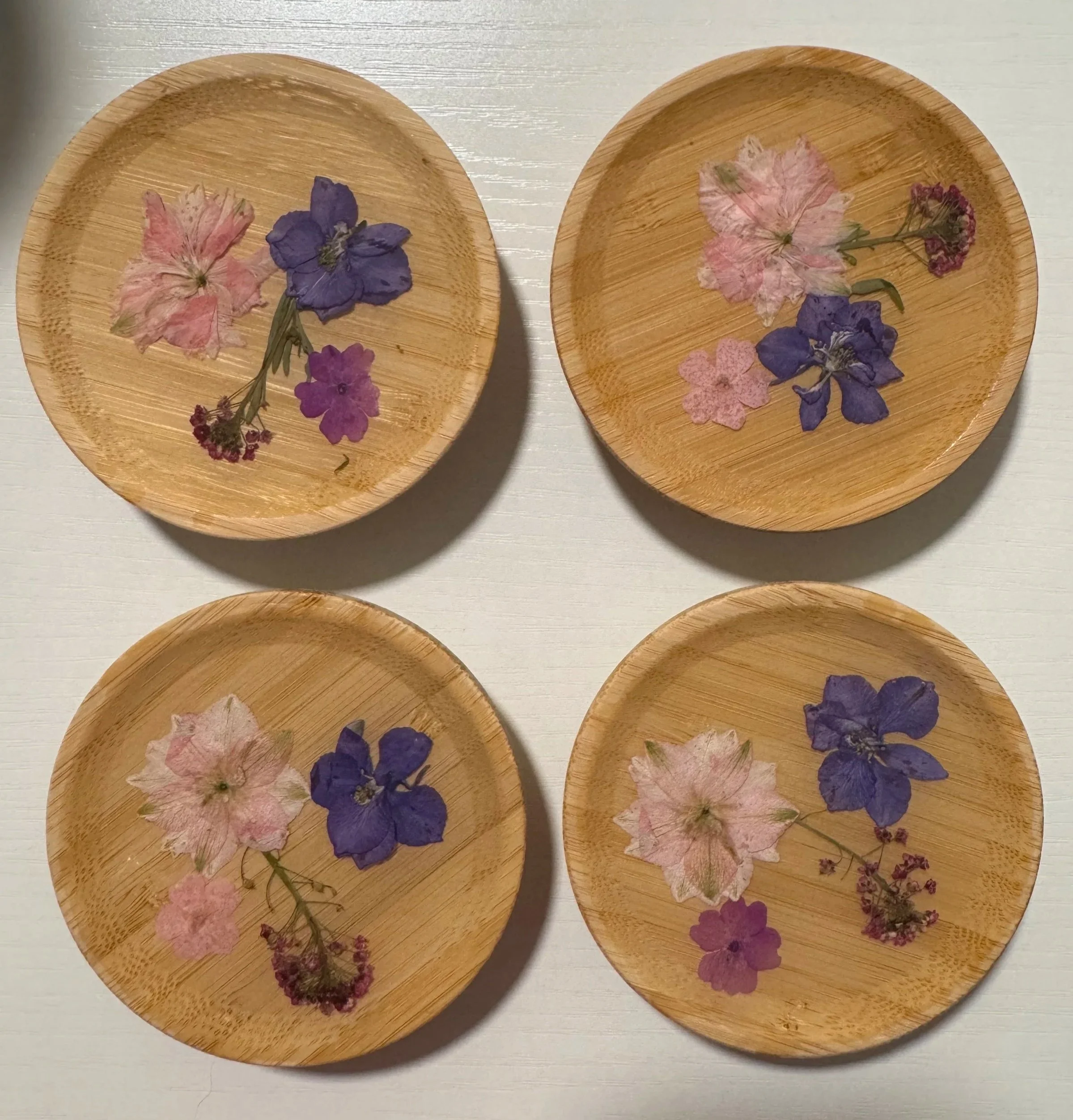 Four circular wooden plates with pressed flowers, including pink and purple blooms, arranged on a light-colored surface.