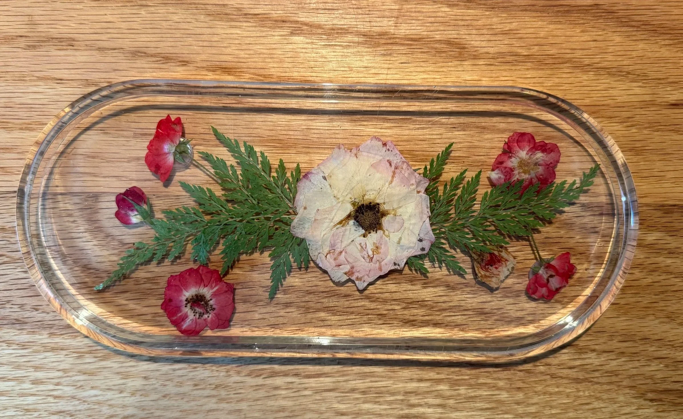 A clear glass tray with a dried flower, a dried leaf, and several small red and pink flowers on a wooden surface.