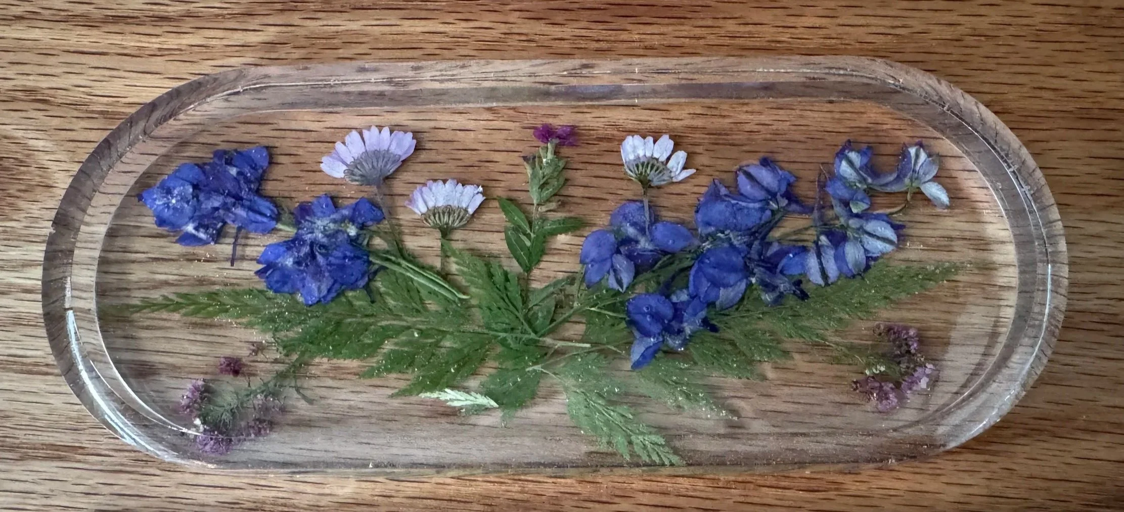 Pressed blue and purple flowers with green leaves inside a clear oval glass tray on a wooden surface.