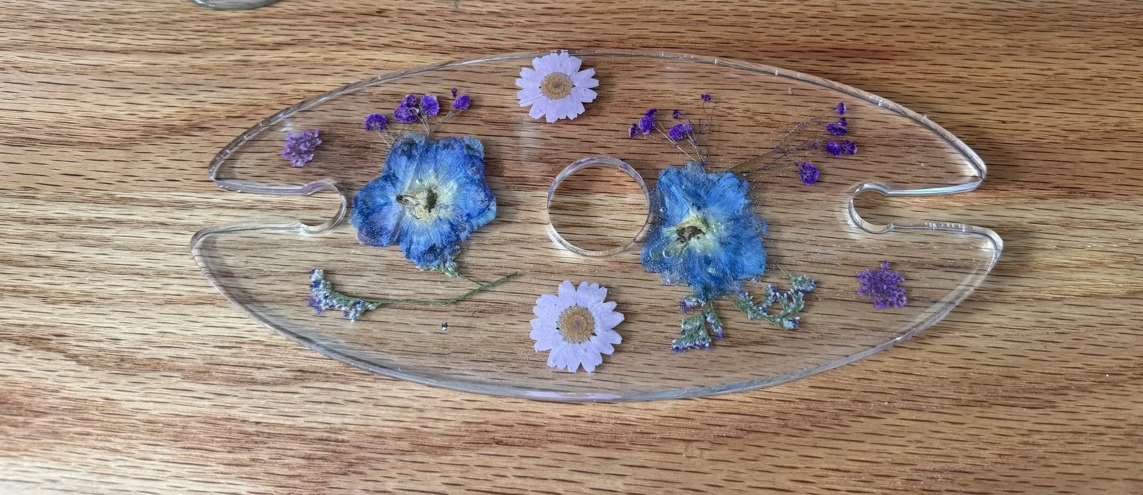 Clear glass eye-shaped tray with pressed dried flowers, including blue pansies, white daisies, and small purple flowers, on a wooden surface.
