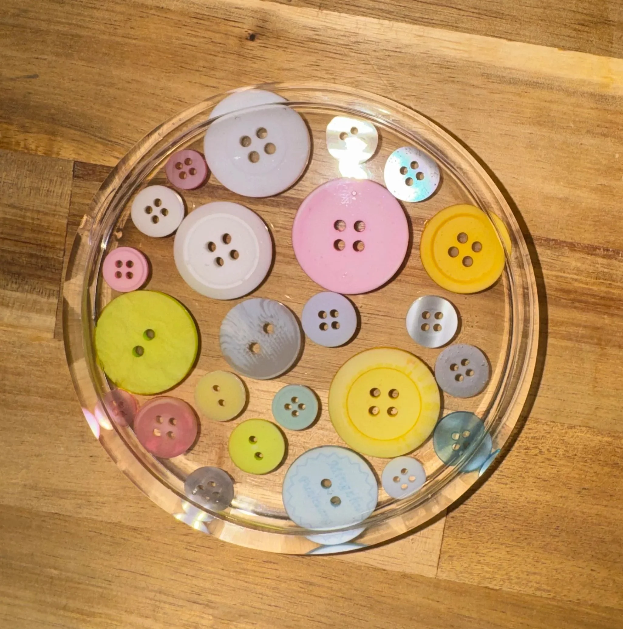 A glass bowl containing various buttons of different sizes, colors (white, pink, yellow, blue, and gray), and designs, placed on a wooden surface.