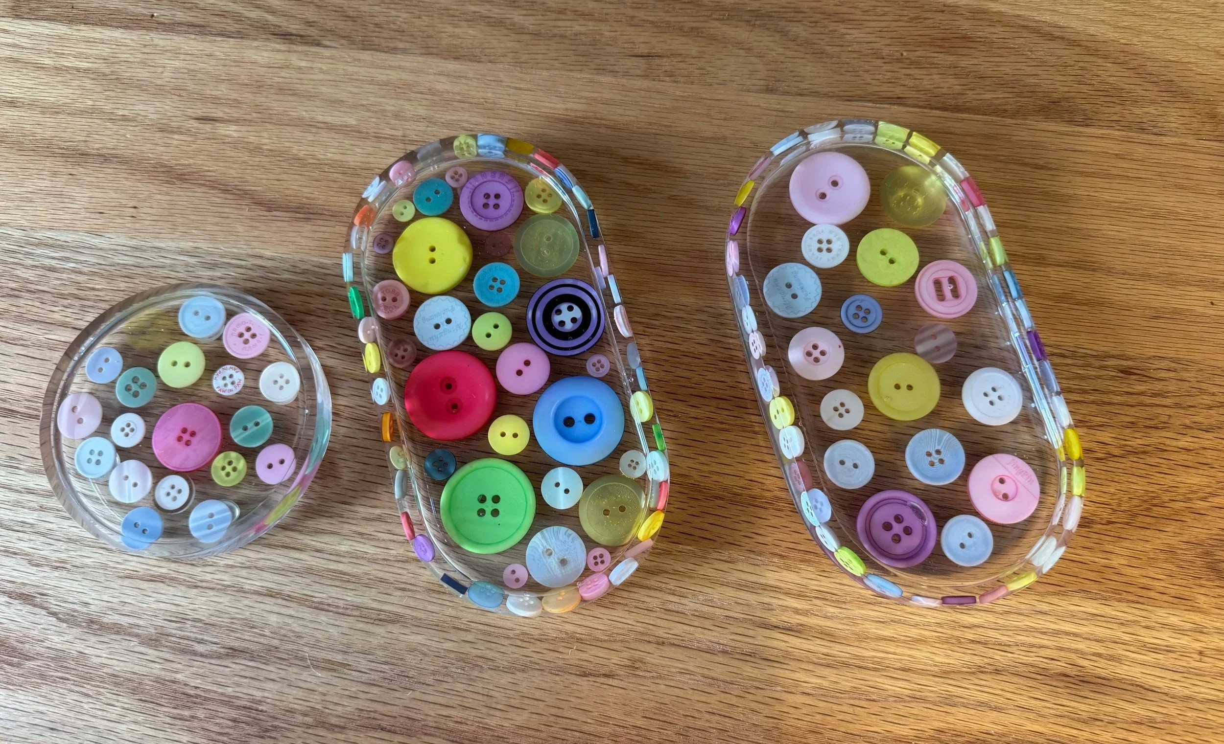 Three clear plastic oval trays filled with colorful buttons of various sizes placed on a wooden surface.