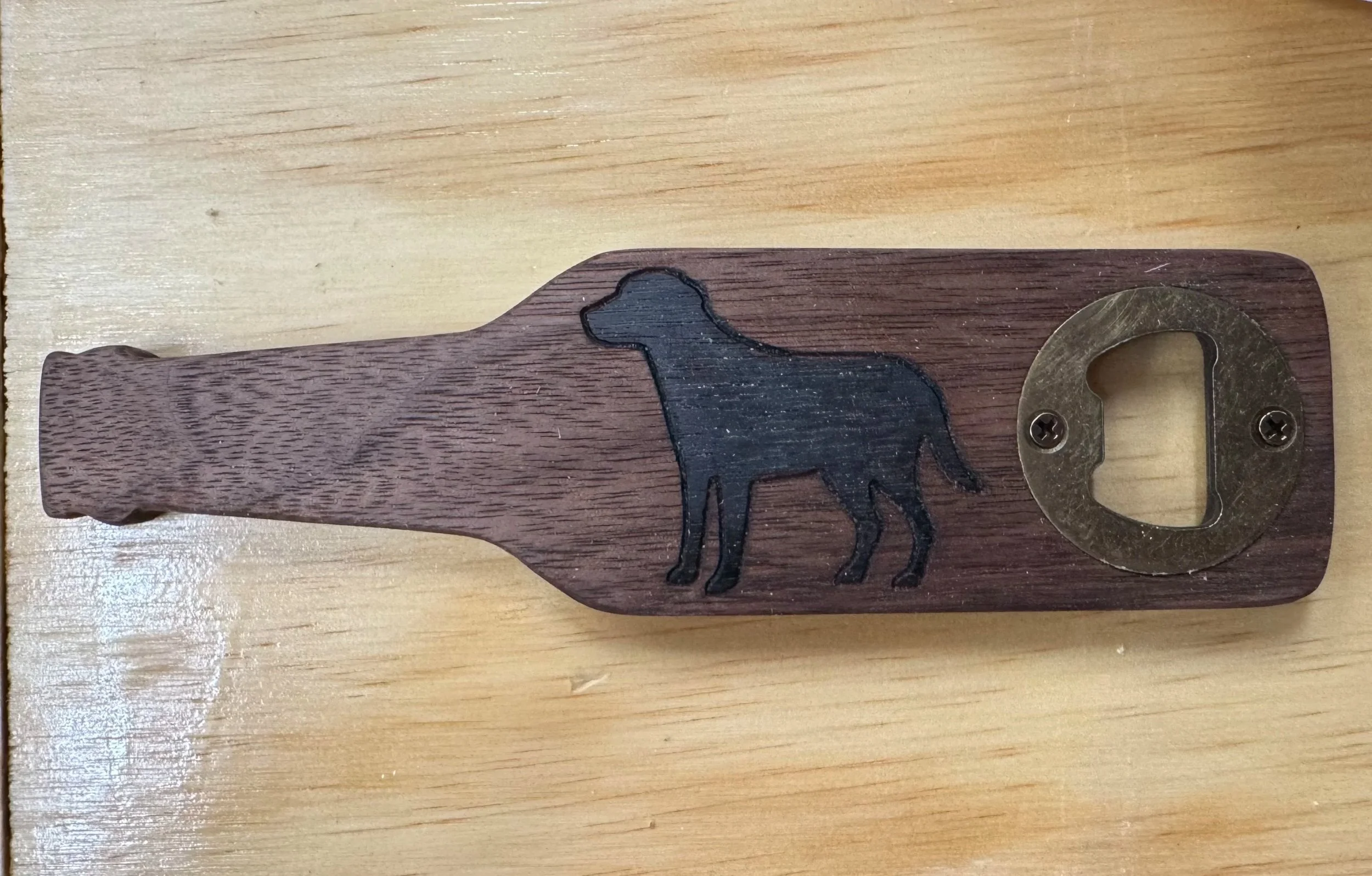 A wooden bottle opener with a black silhouette of a dog carved into it and a metal bottle opener attached to the right side, placed on a light-colored wooden surface.