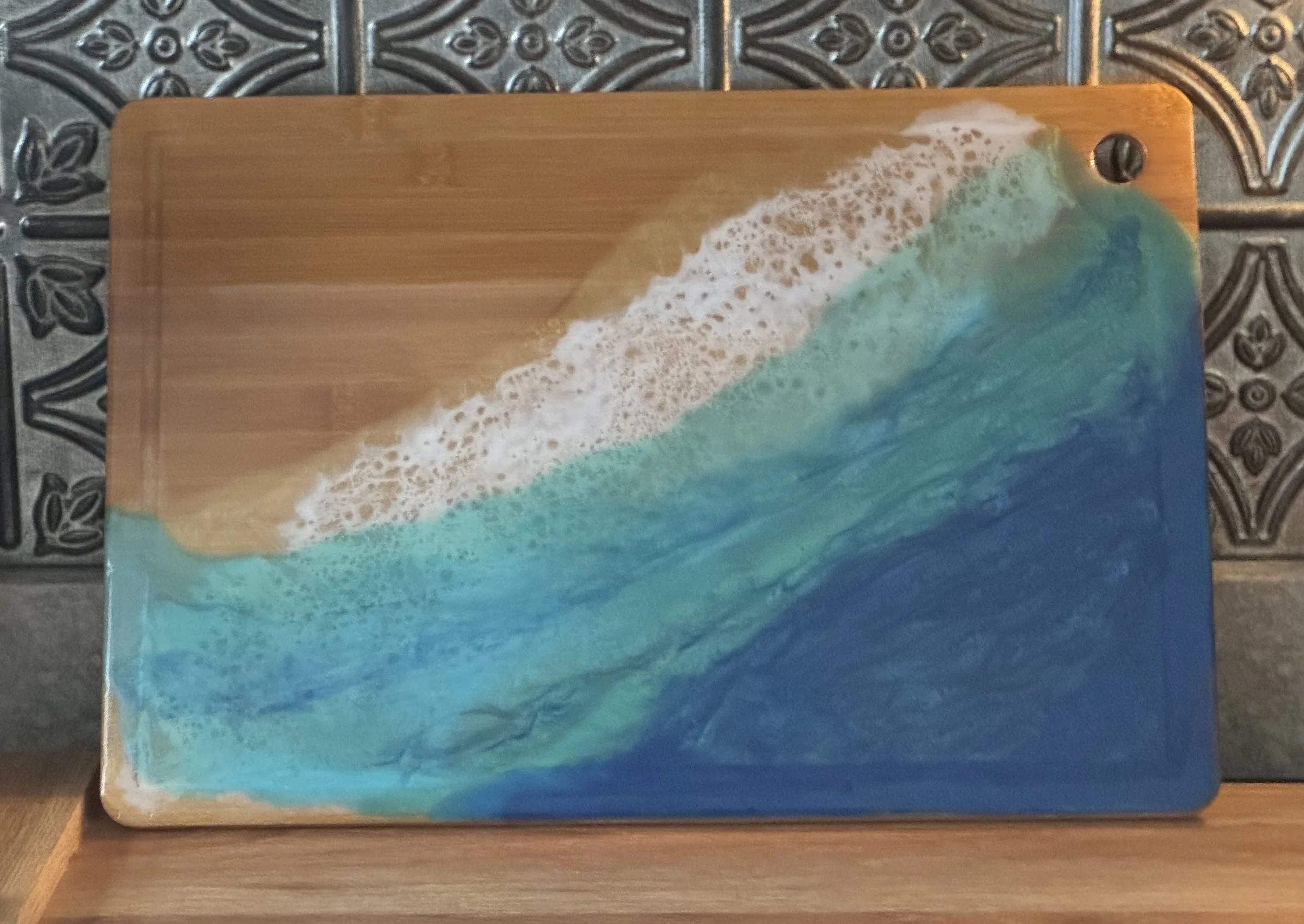A wooden cutting board with a design of a beach scene, featuring sand, waves, and foamy ocean water, placed on a dark patterned tile surface.