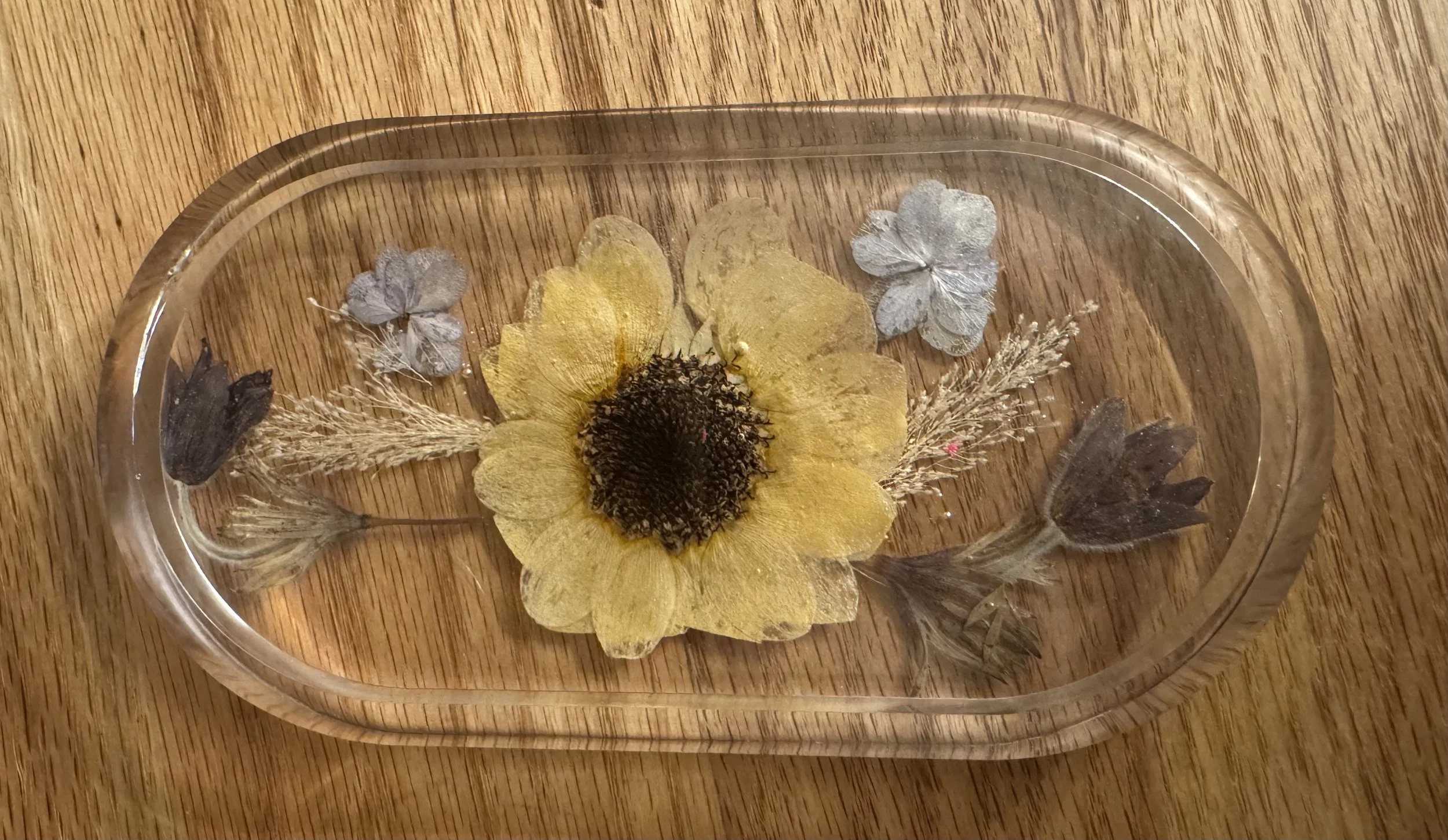 A glass dish with a yellow flower, two small grey flowers, and some dried plant elements on a wooden surface.
