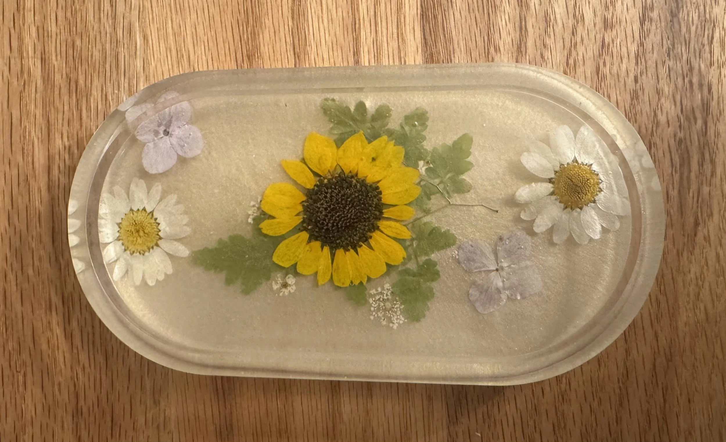 Pressed flower arrangement of daisies and a sunflower in a glass container on a wooden surface.