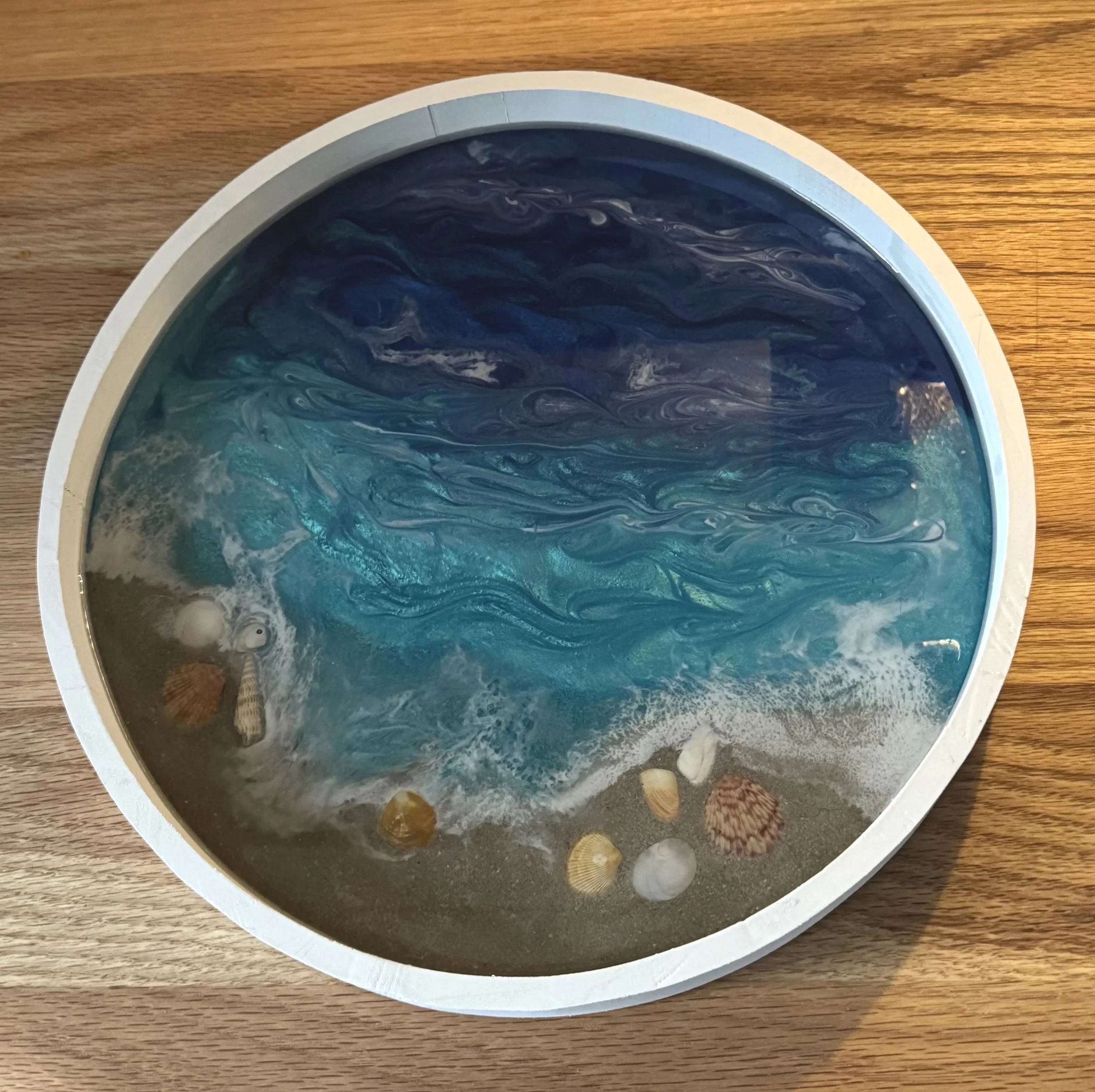 An overhead view of a round, marble-patterned tabletop display with an ocean scene in a container, showing various seashells at the bottom edge.