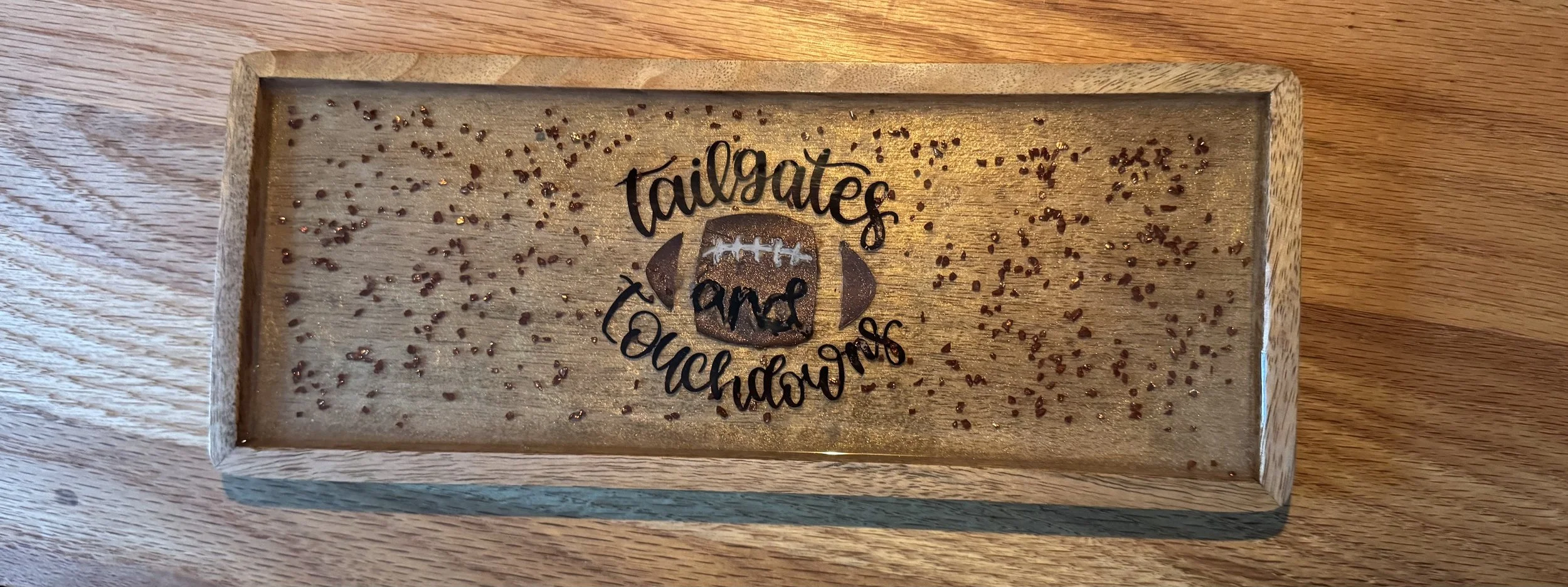 A wooden rectangular tray with gold glitter and small chocolate sprinkles, featuring a black decal of a football with the words "tailgate and touchdowns" around it.