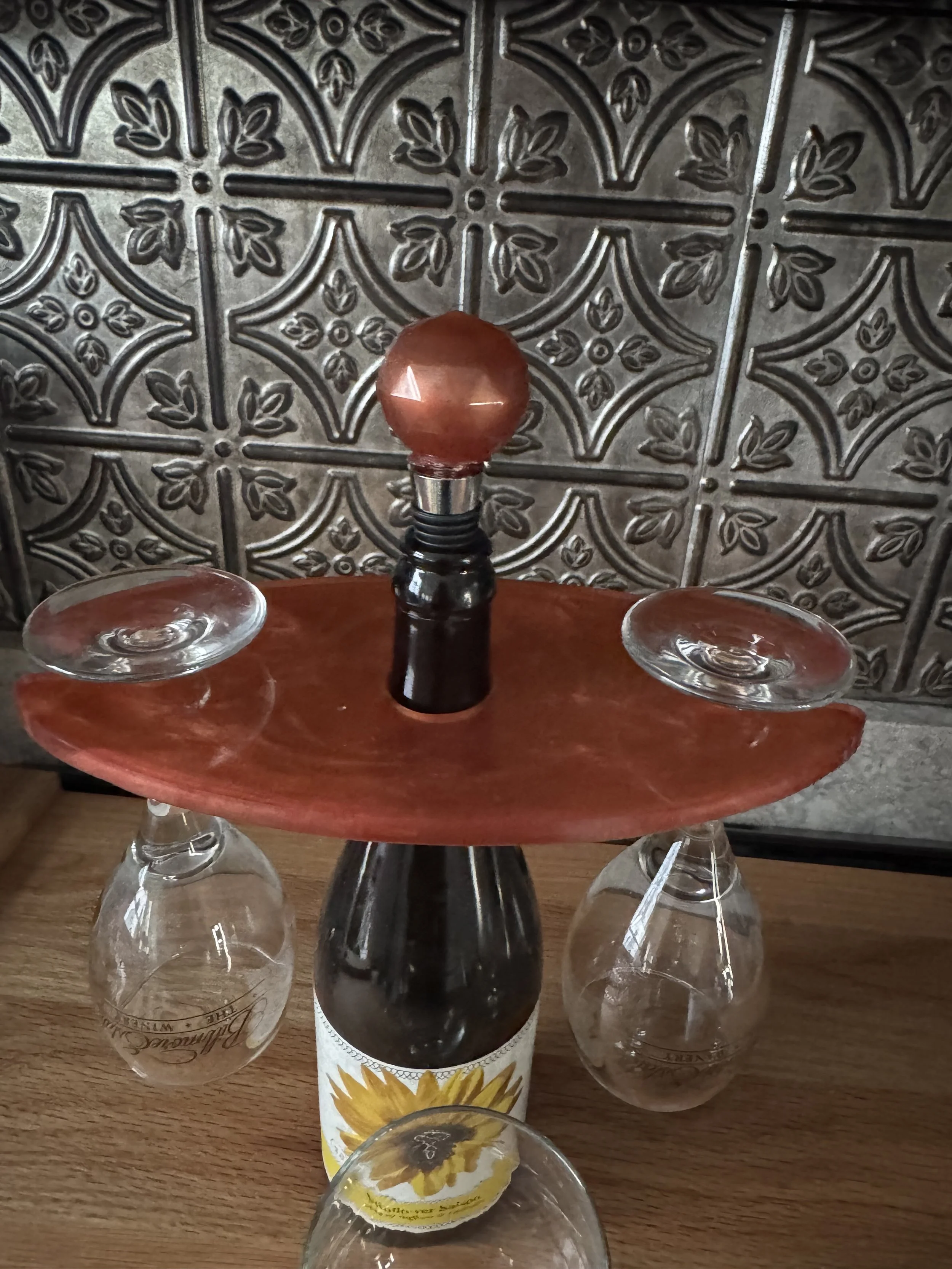 A wine bottle with a sunflower illustration on the label, placed on a wooden surface, surrounded by upside-down wine glasses, with a textured decorative wall in the background.