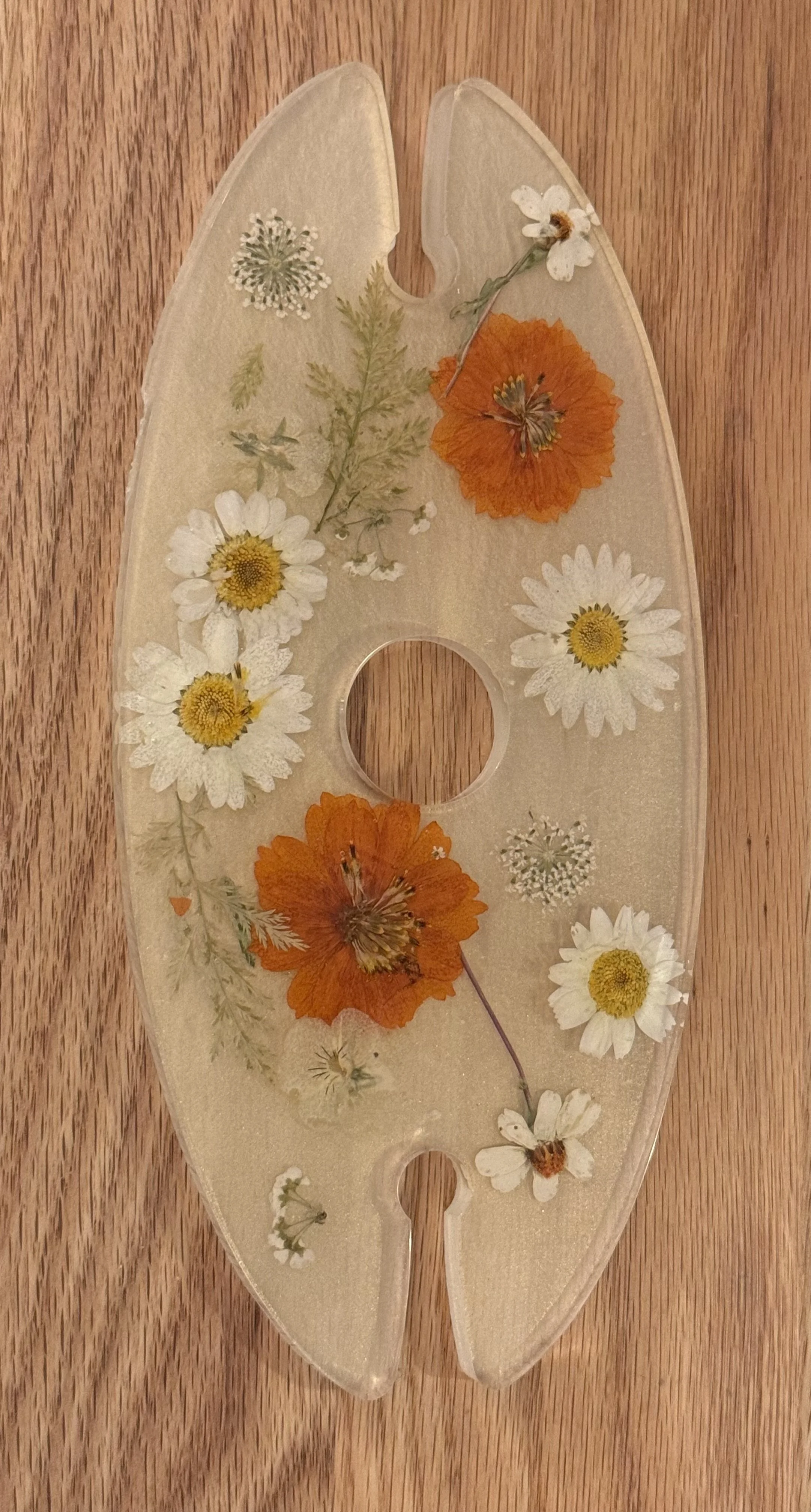 A transparent oval glass palette decorated with pressed flowers and herbs, resting on a wooden surface.