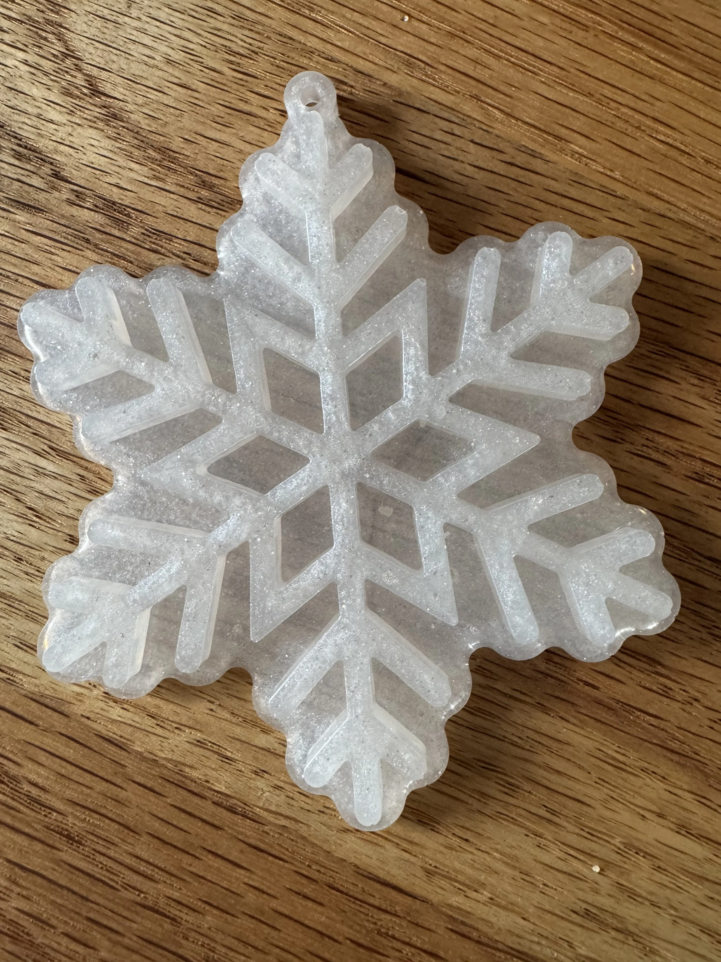 White shimmery resin snowflake ornament/decoration with intricate design, resting on wooden surface.