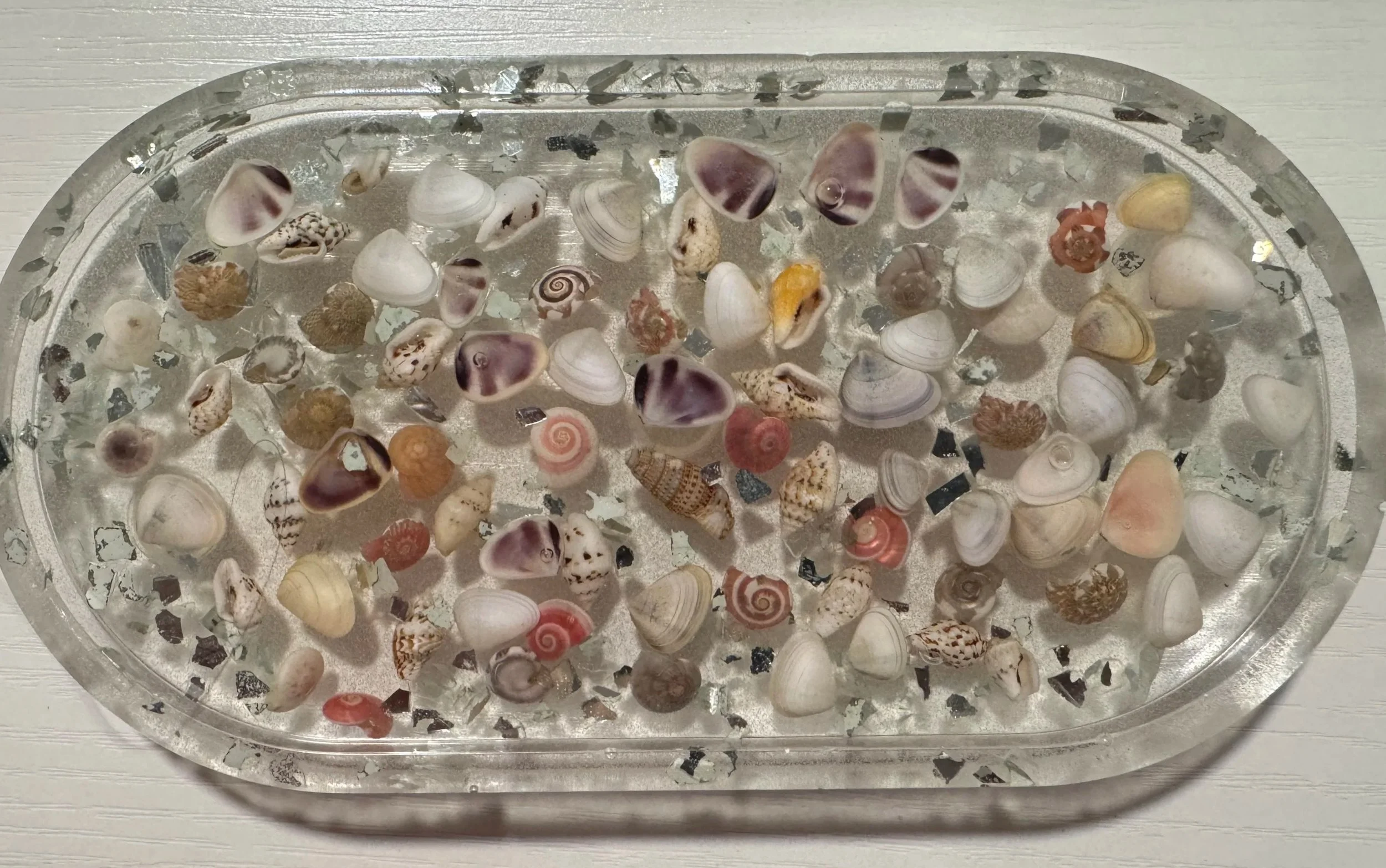 A silver, oval-shaped tray filled with various small seashells of different colors and patterns, placed on a light-colored wooden surface.