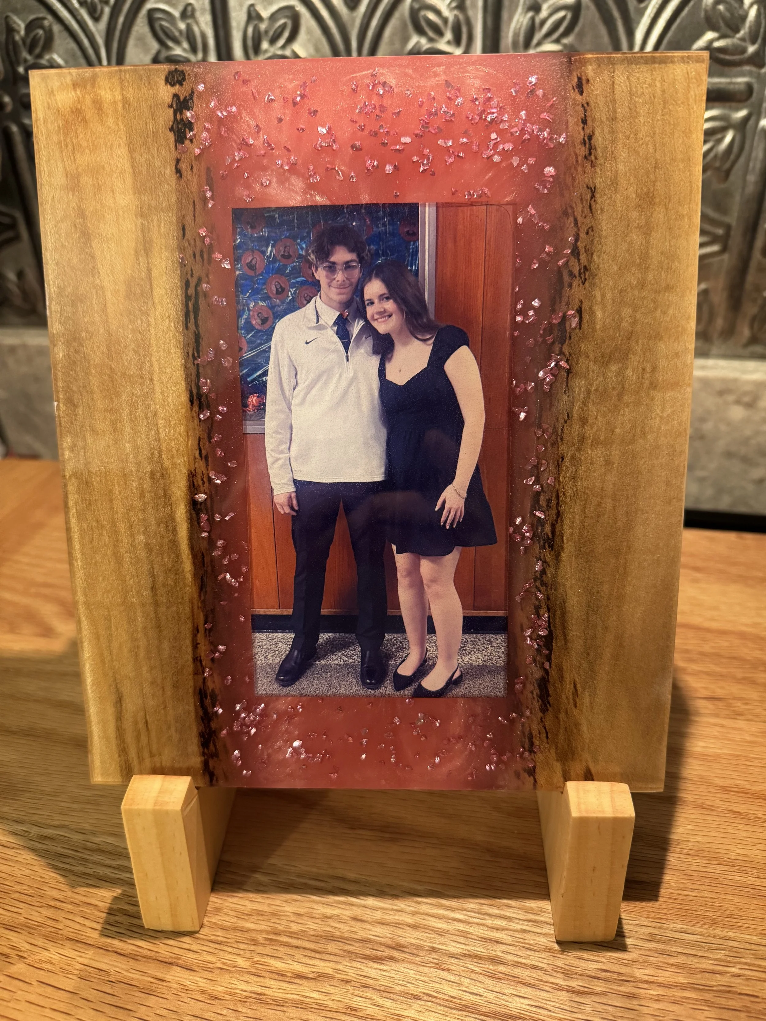 A wood and resin photo frame holding a picture of a young man and woman embedded in resin. The frame has a wooden outer part with pinkish-red accents and small decorative elements.