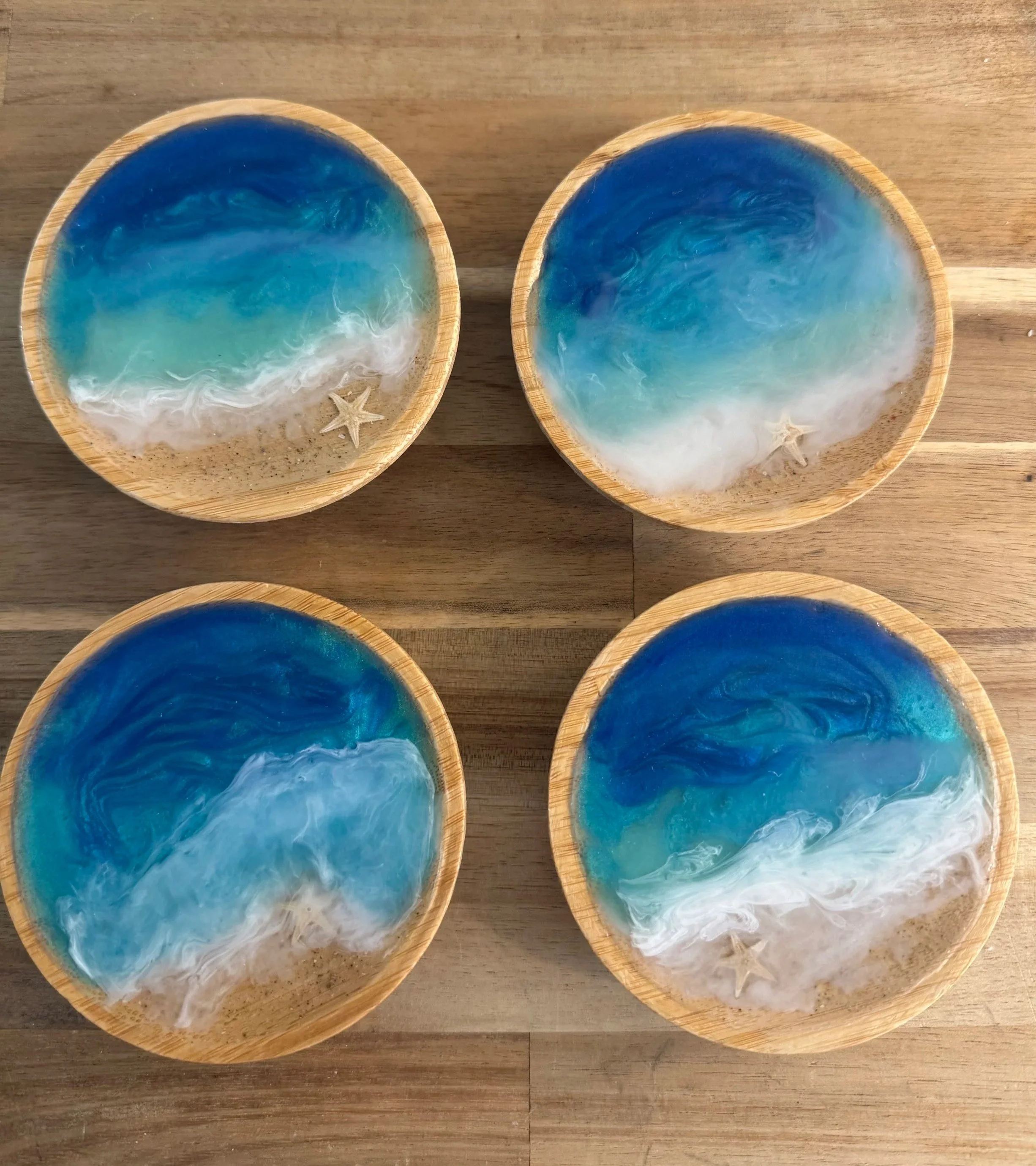 Four small wooden bowls with ocean wave designs, filled with a mixture of clear and blue resin to resemble the ocean, with starfish decorations inside the bowls, arranged on a wooden surface.